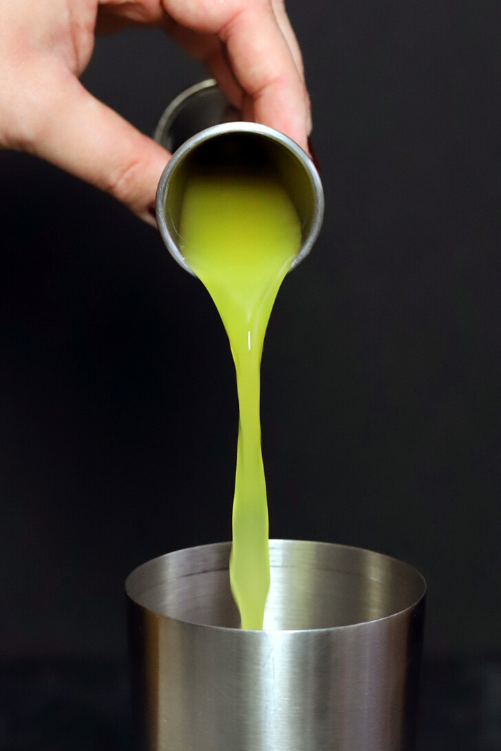 A hand pouring orange juice from a jigger into a cocktail shaker.
