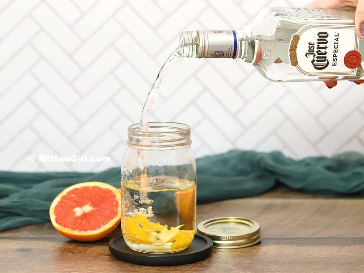 Tequila being poured into a pint jar with grapefruit peels. 