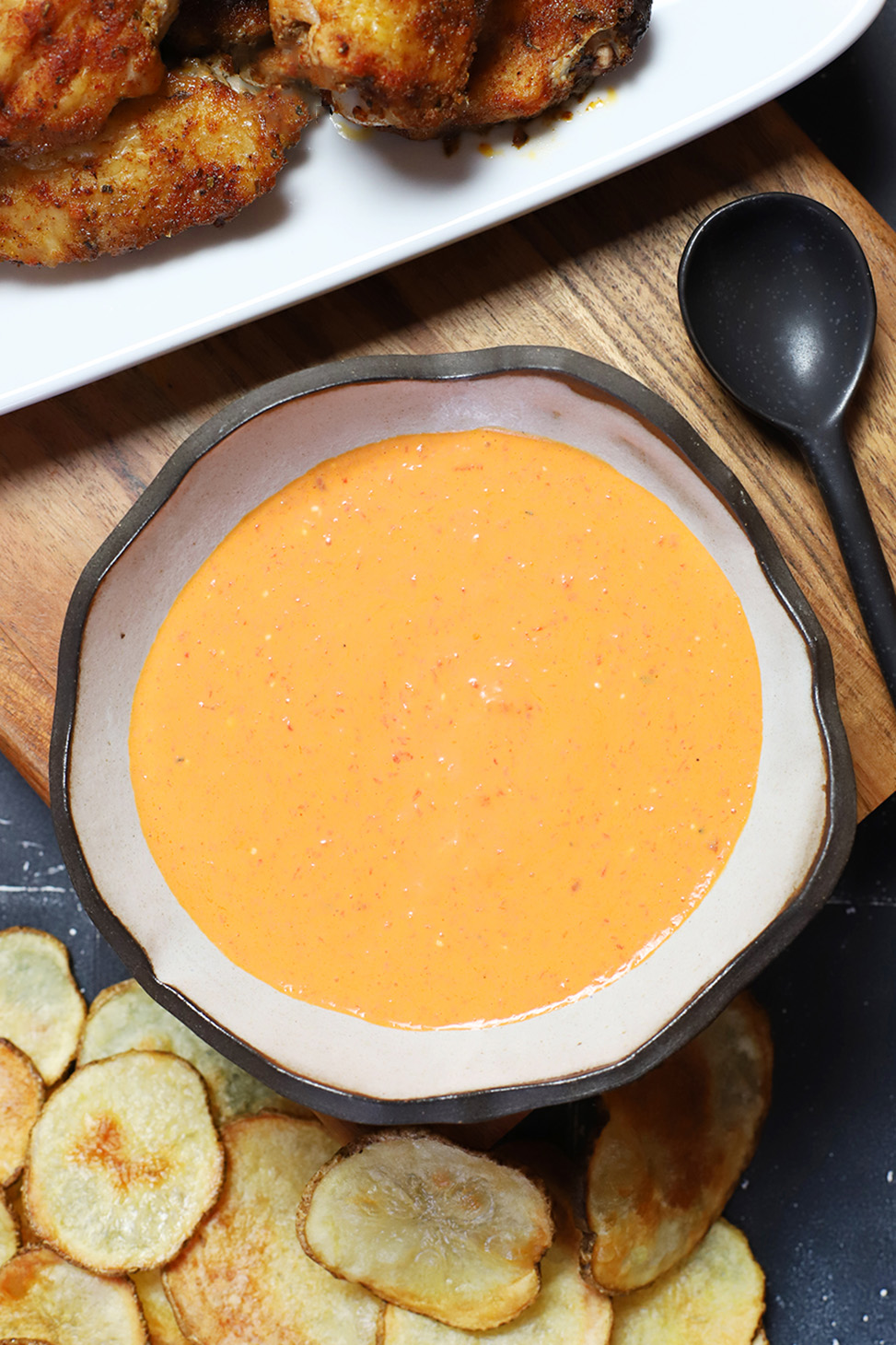 A bowl of roasted red pepper and goat cheese dip next to a spoon, some potato chips and a platter of chicken wings. 