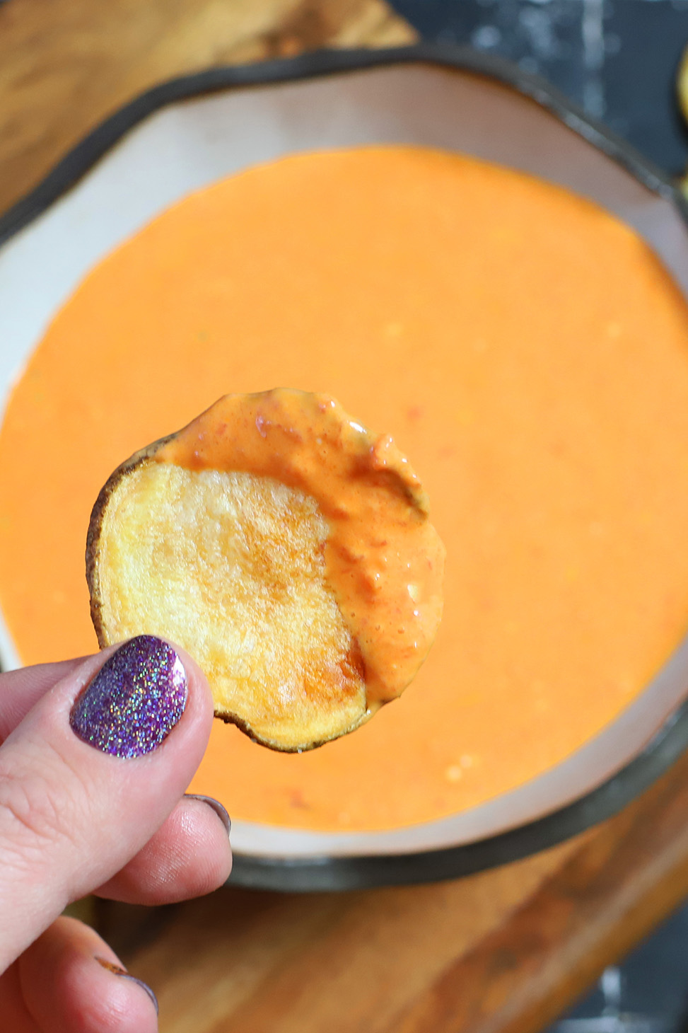A hand holding a chip that has been dipped in roasted red pepper and goat cheese dip over a bowl of the same dip.