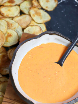 A bowl of roasted red pepper and goat cheese dip with a spoon in it next to some potato chips.