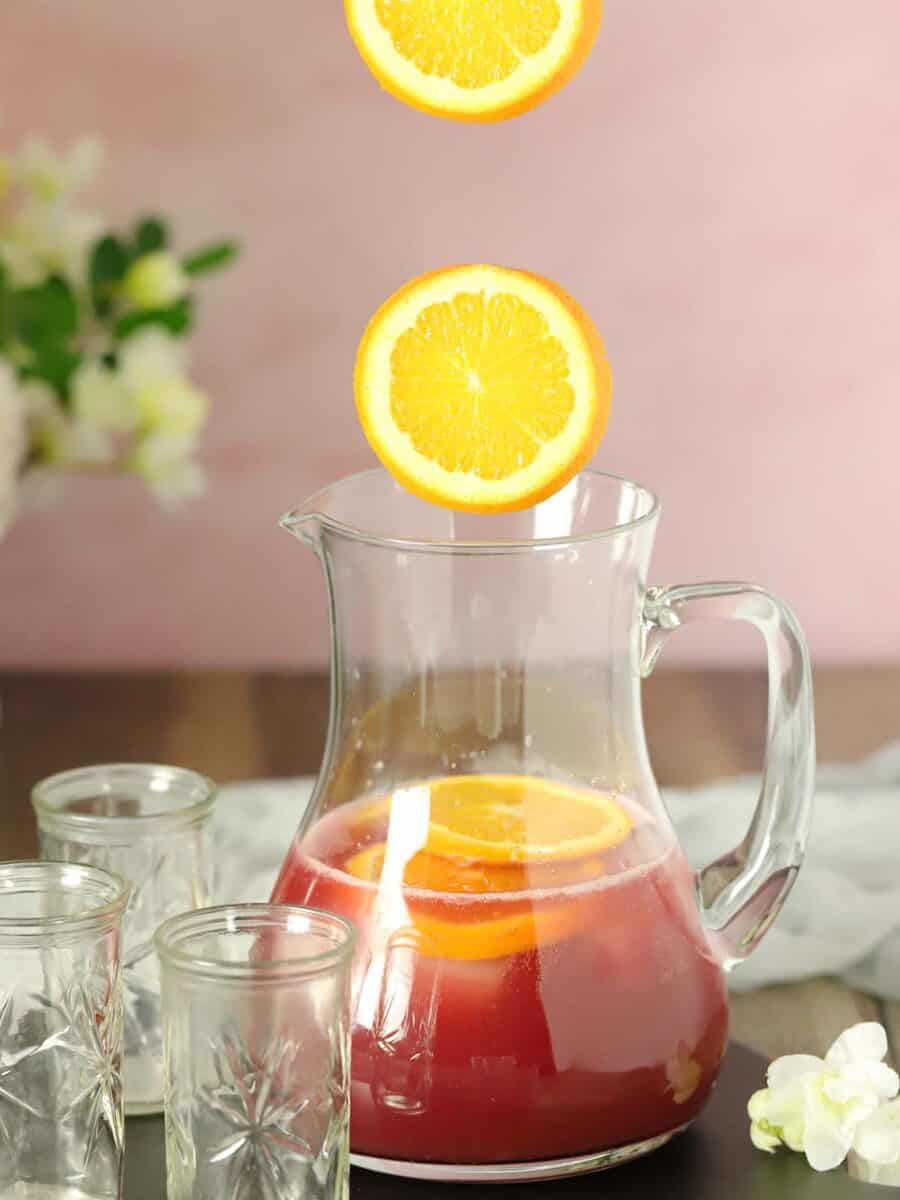 Sliced oranges being dropped into a pitcher of red sangria mocktails