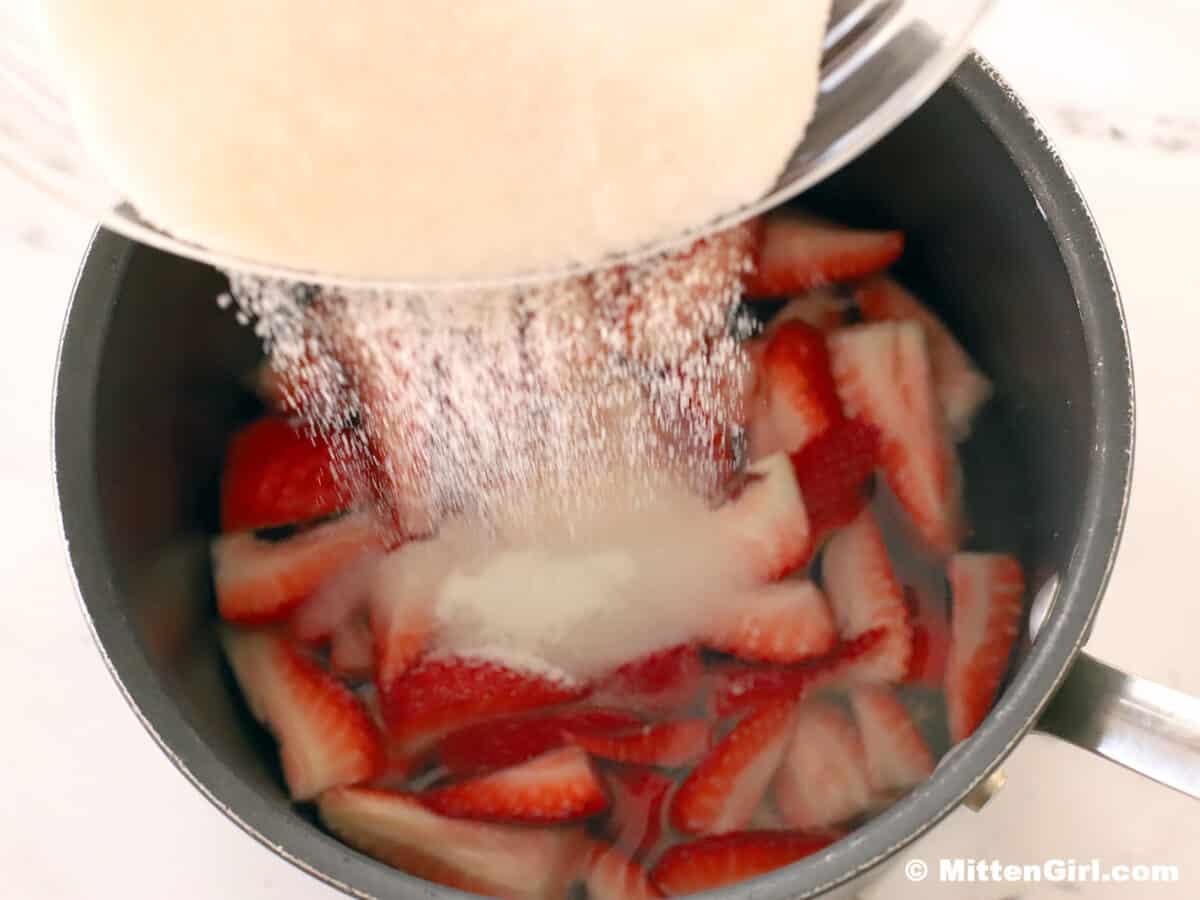 Sugar being poured into a pot of sliced strawberries and water.