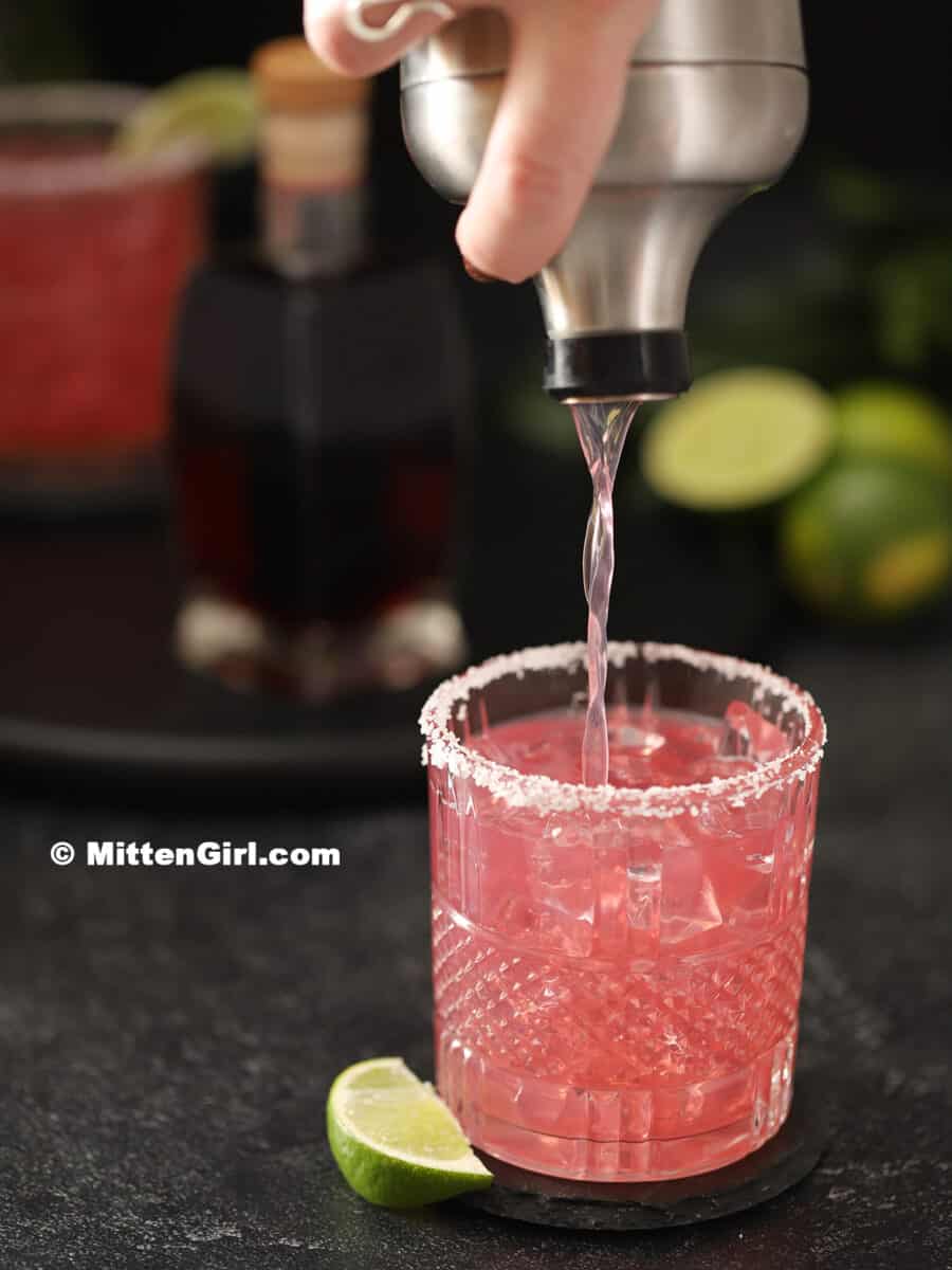 Pouring a hibiscus margarita from a cocktail shaker into a glass. 