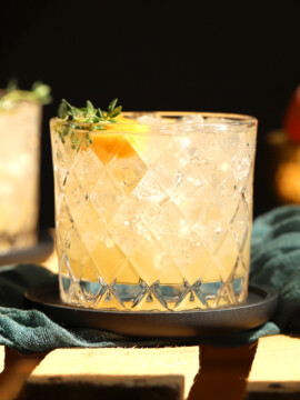 A glass of non alcoholic peach sangria sitting in the sunlight, garnished with a slice or peach and sprig of thyme.