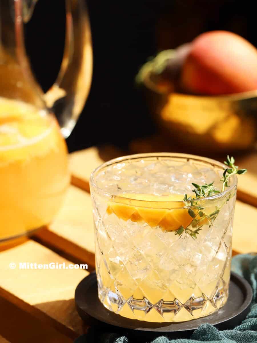 A glass of non alcoholic peach sangria sitting in the sunlight, garnished with a slice or peach and sprig of thyme.