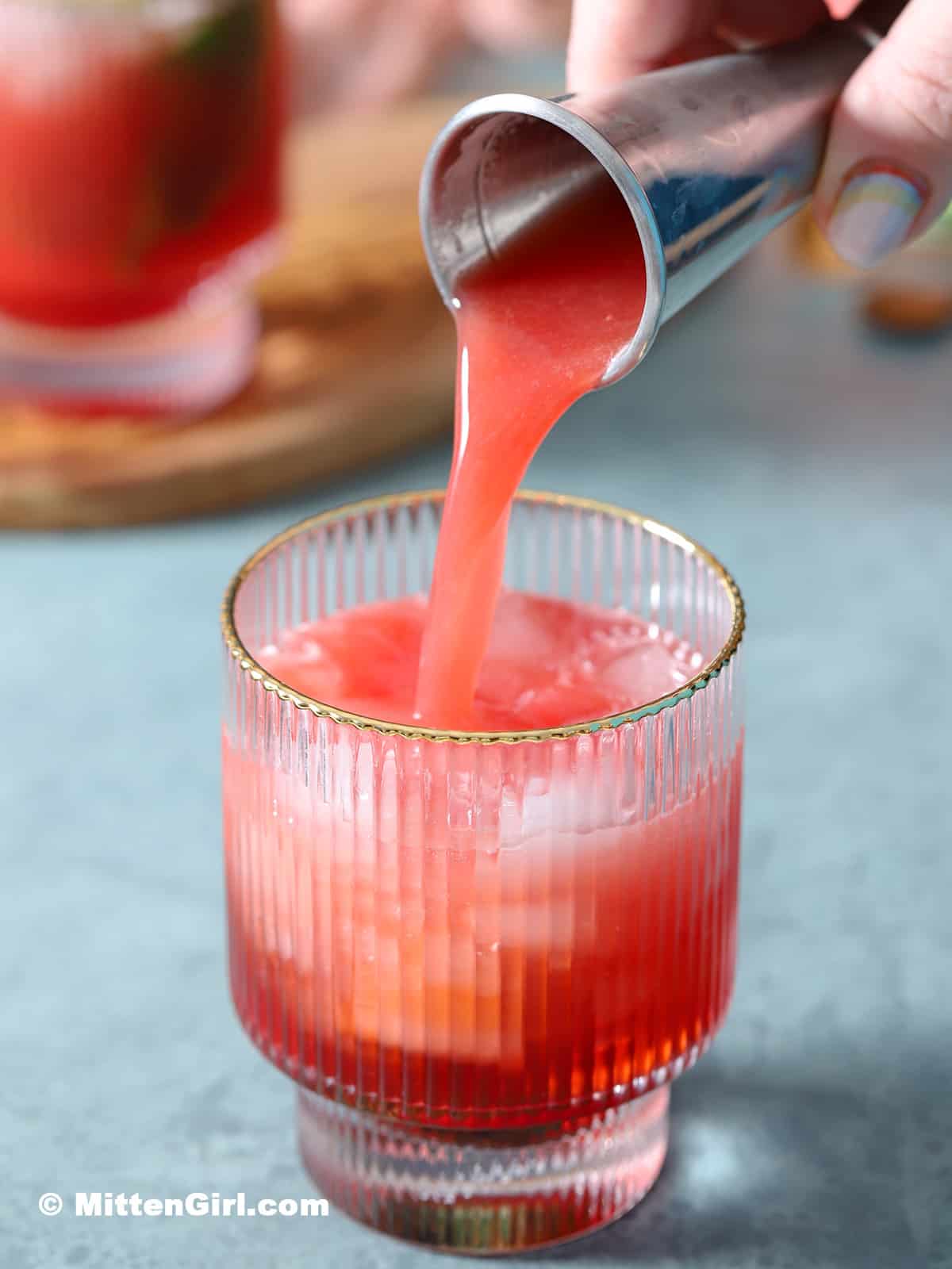 Pouring watermelon juice into a glass filled with ice, mint, lime juice and strawberry syrup. 