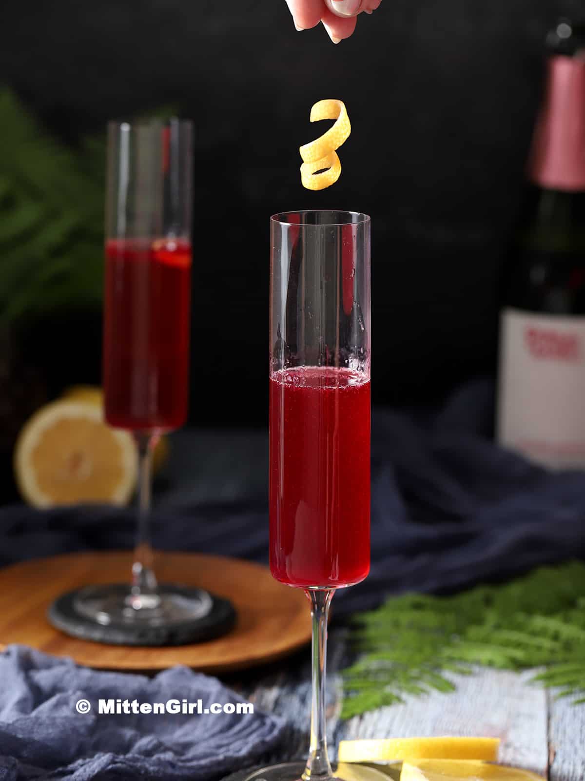 A lemon peel twist being dropped into a glass of blueberry french 75 cocktail.