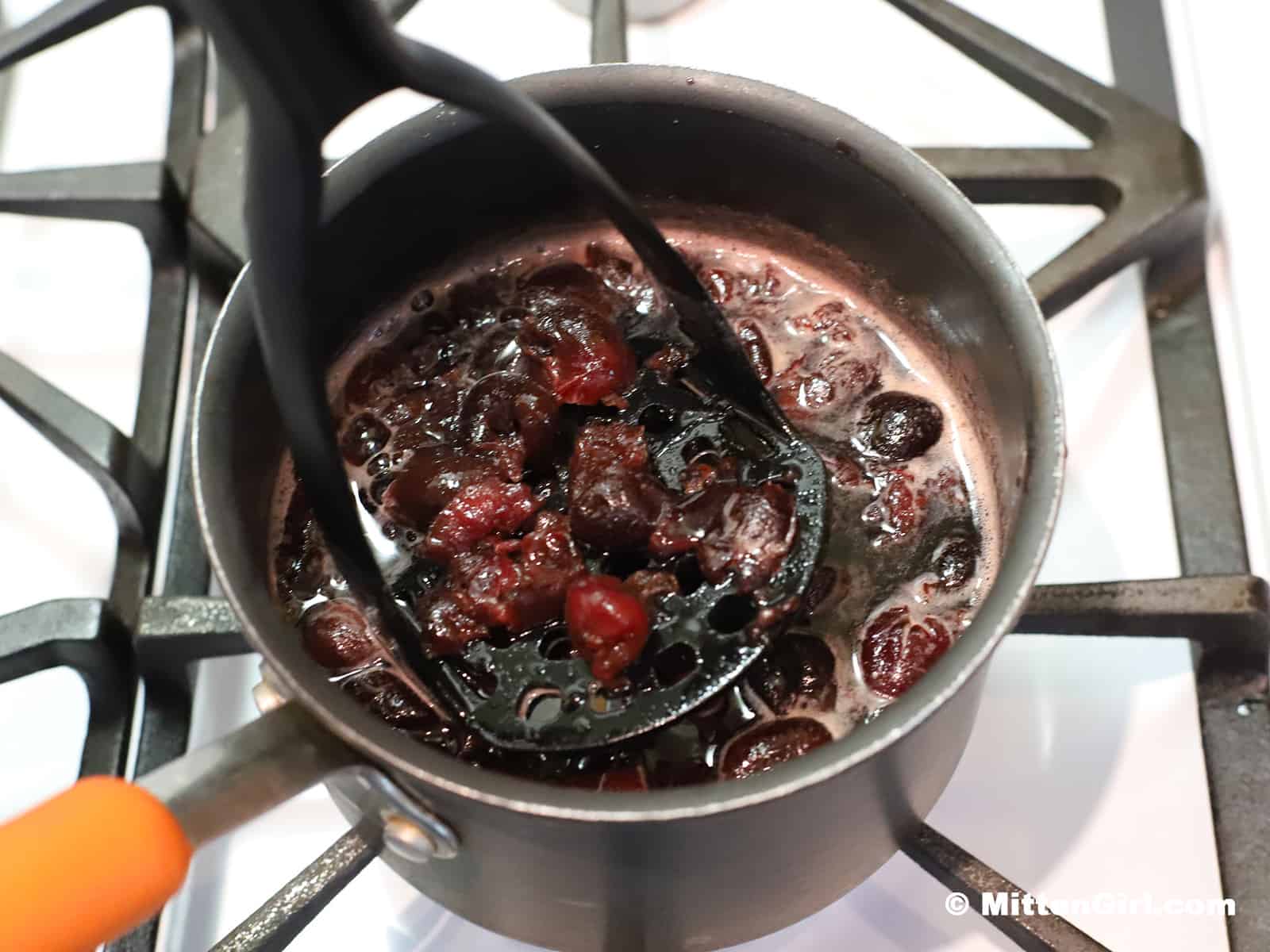 Cooked cherries being mashed in a pot with a potato masher. 