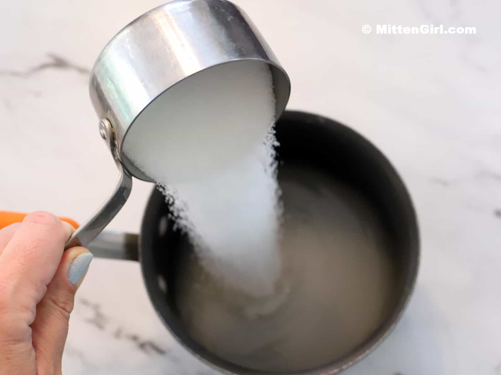 A cup of sugar being poured into a pot of water. 