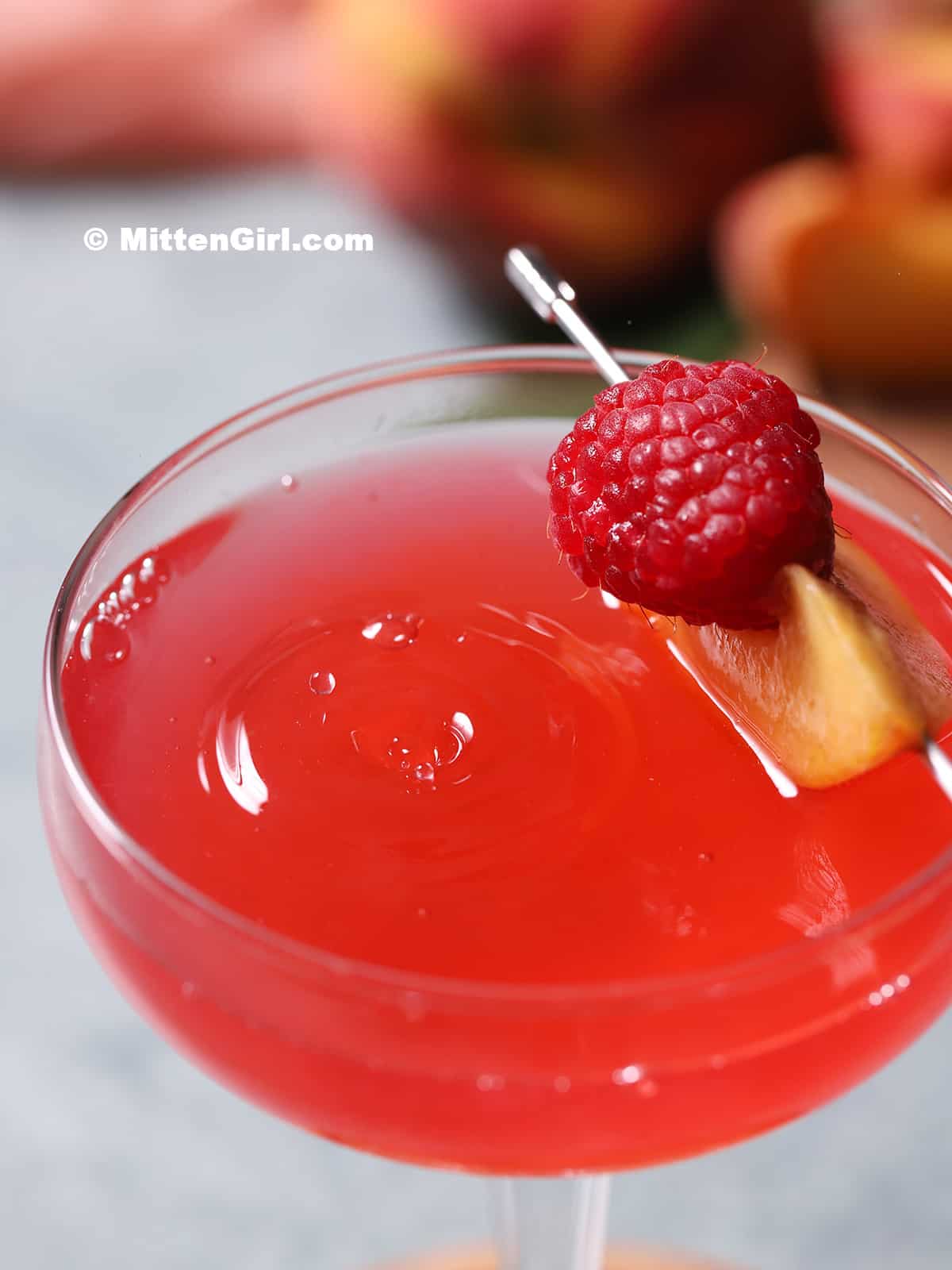 A coupe glass filled with peach raspberry martini, garnished with a slice of peach and a fresh raspberry.