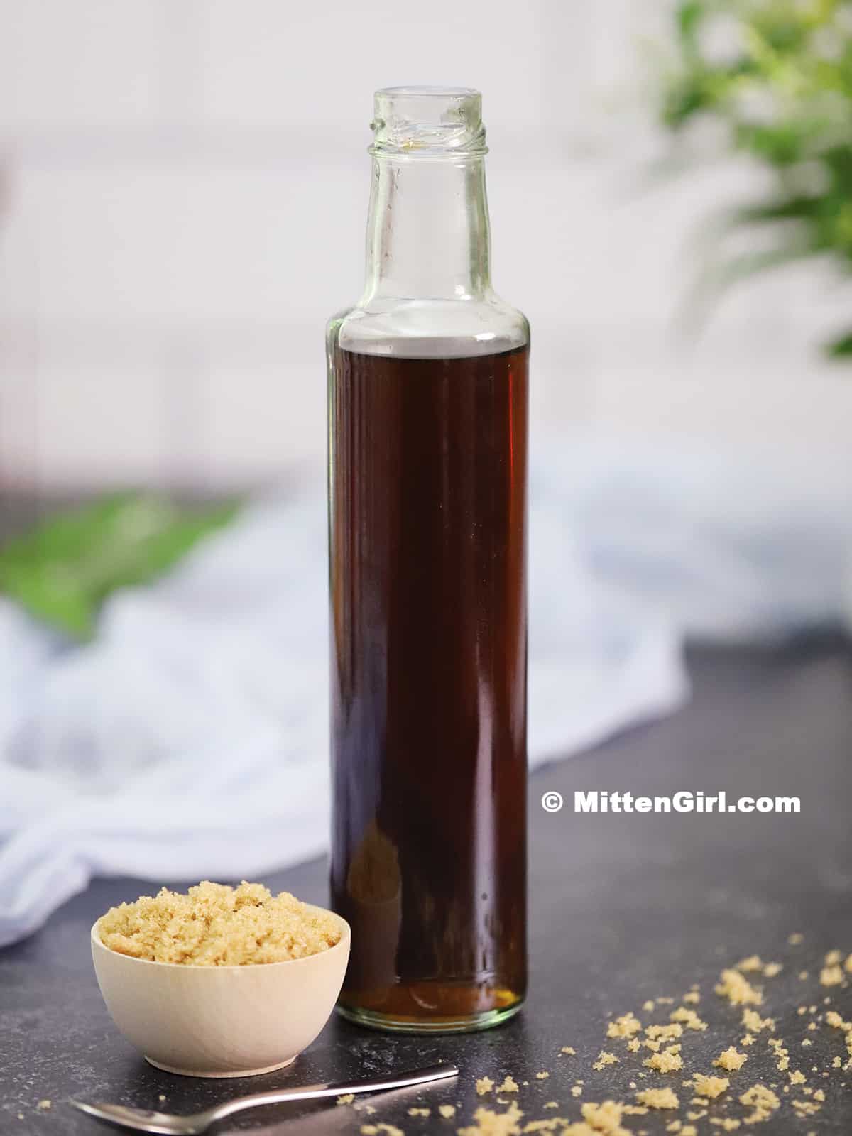 A small bowl of brown sugar next to a tall bottle of brown sugar syrup.