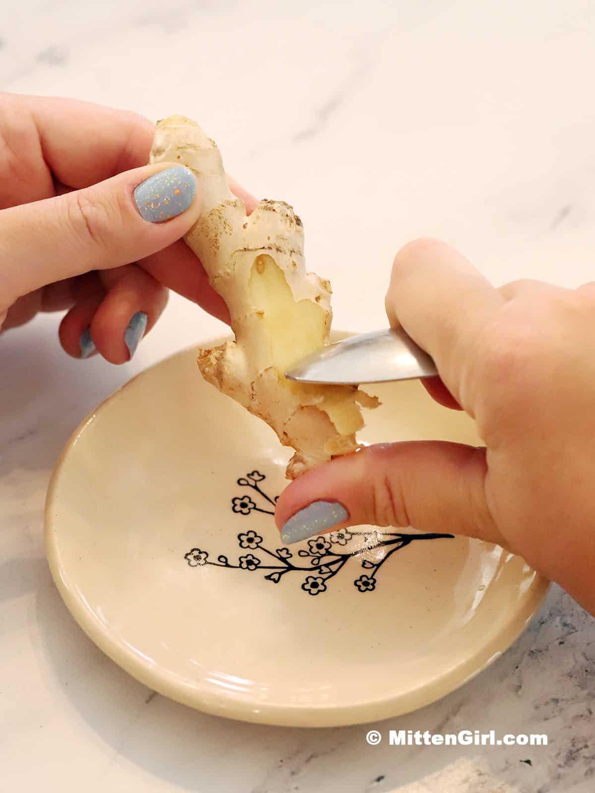 Hands holding a ginger root and using a spoon to peel it.