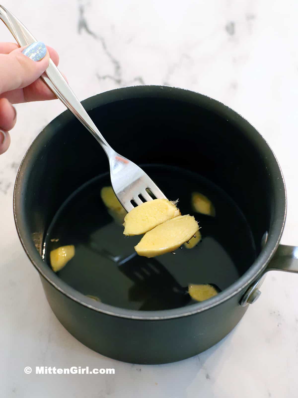 A fork lifting pieces of ginger root out of a pot of syrup.