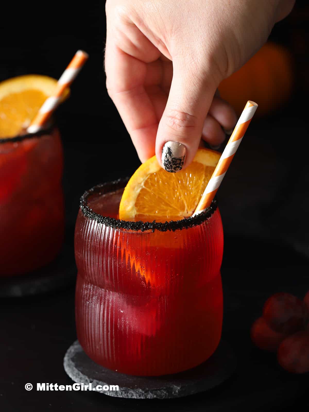 A hand placing a slice of orange into a glass filled with Halloween mocktail.