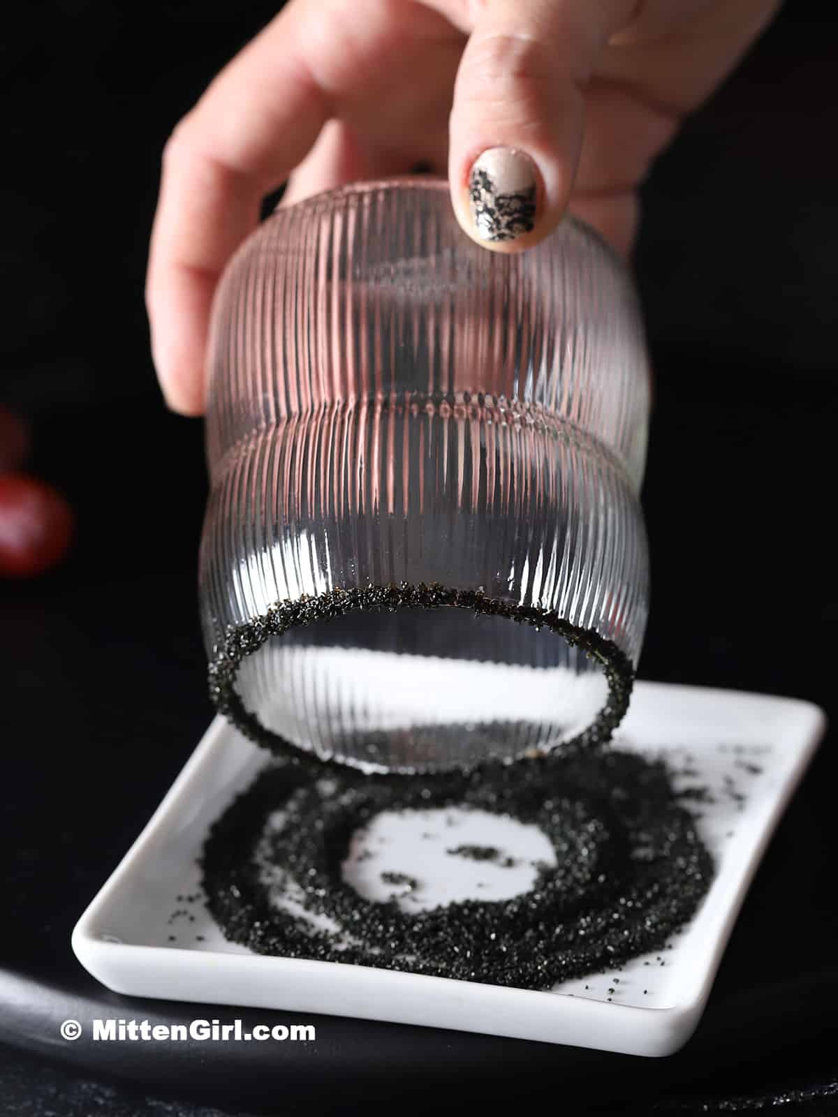 A hand rolling the rim of a glass in a small plate of black sugar.