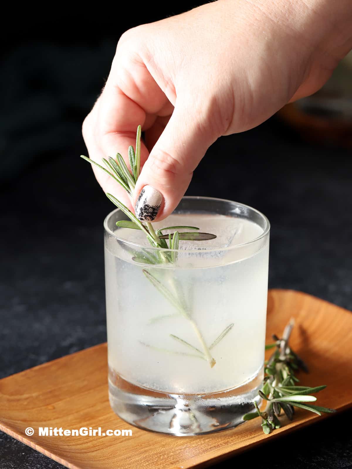 A hand placing a sprig of rosemary into the finished cocktail.