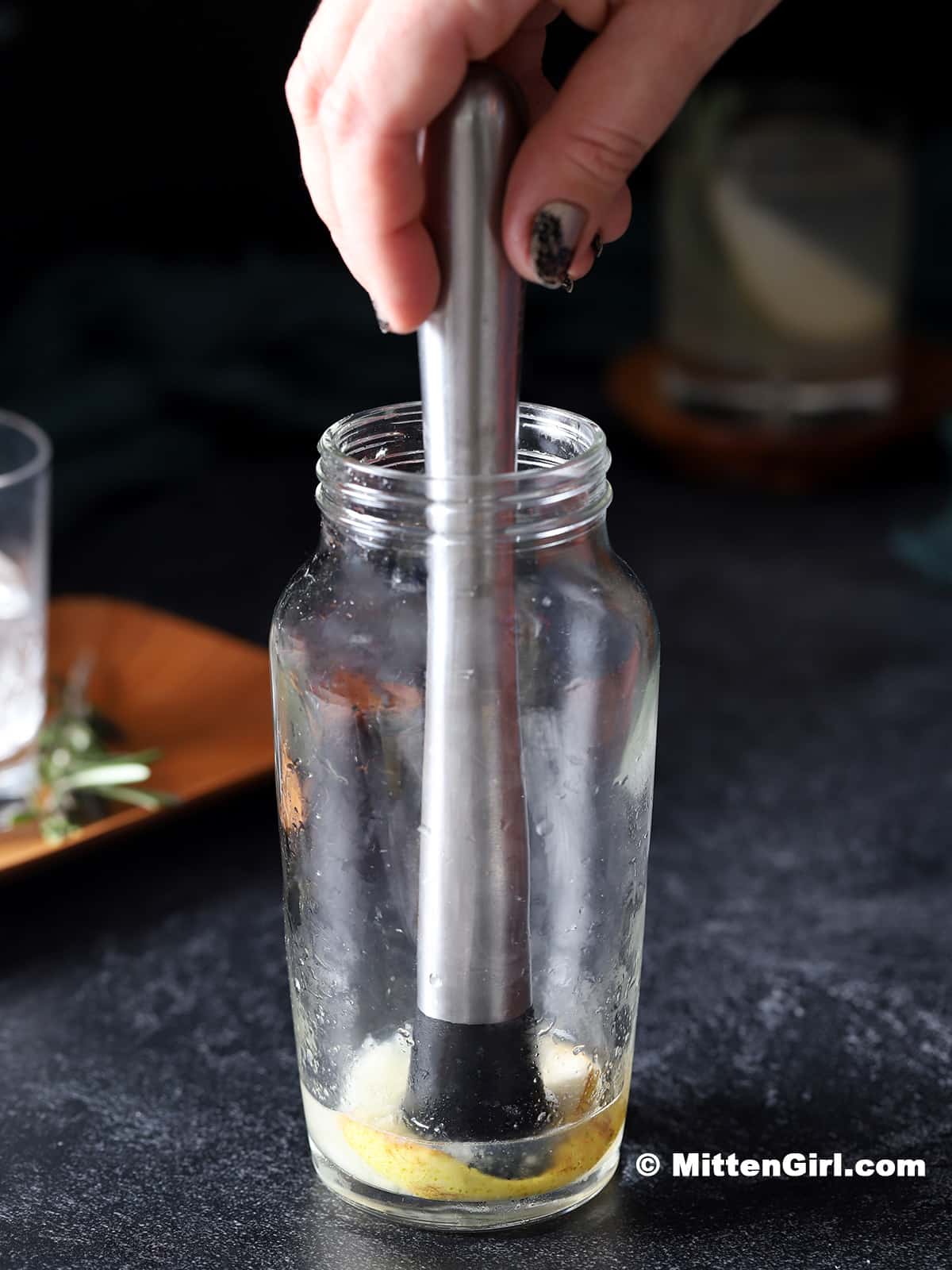 A hand using a muddler to crush a slice of pear in a glass cocktail shaker. 
