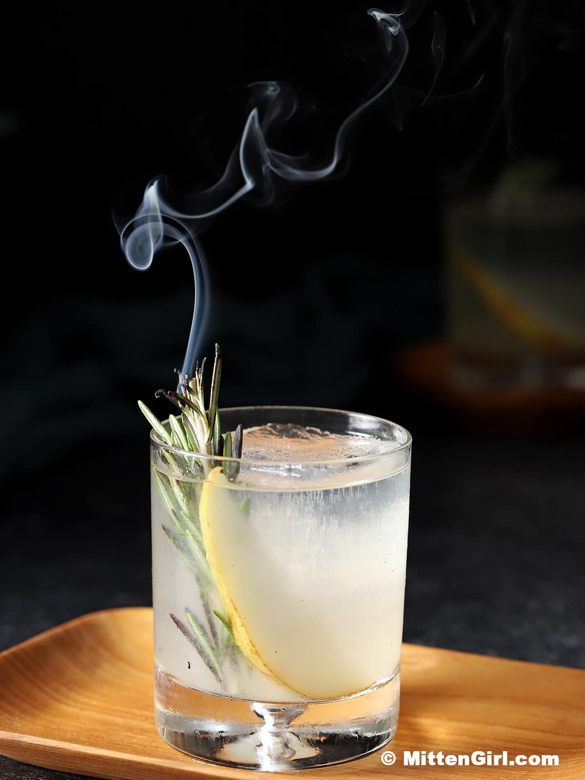 A glass of pear gin cocktail with a piece of rosemary. Smoke is rising from the rosemary.