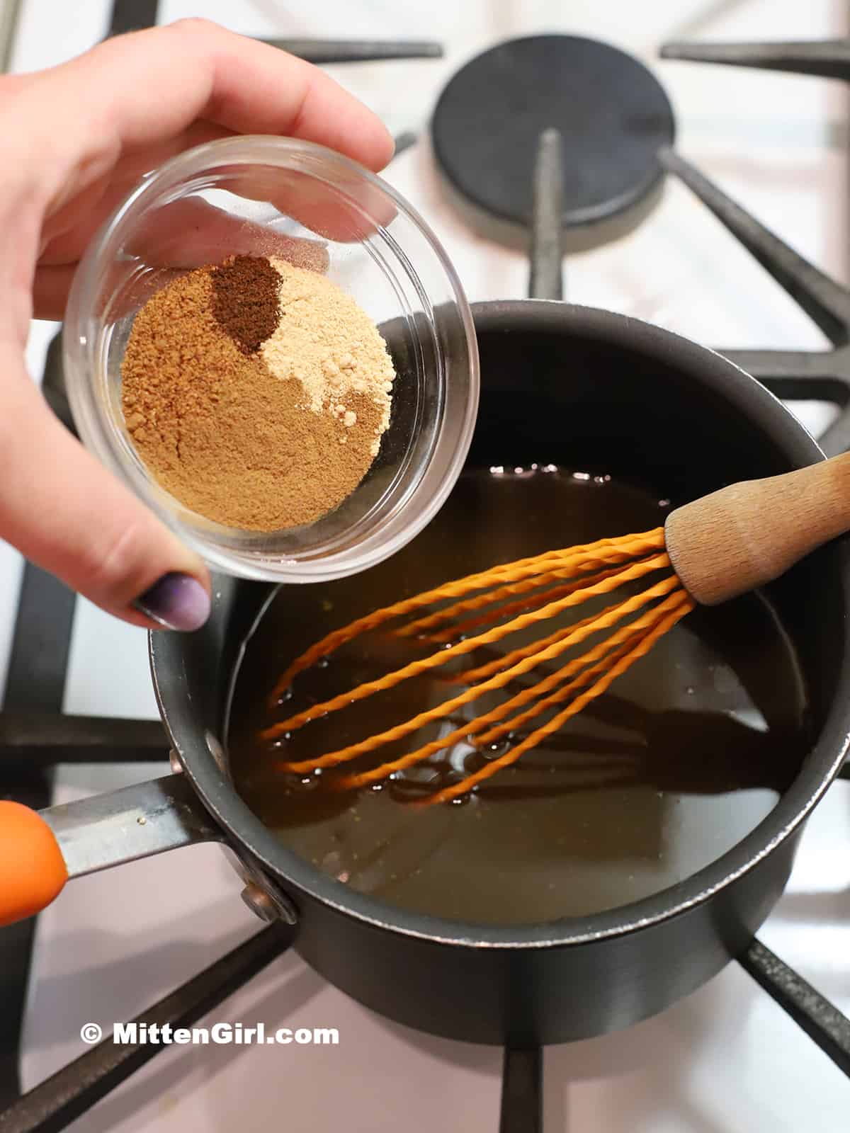 A hand adding spices to a pot of brown sugar syrup. 
