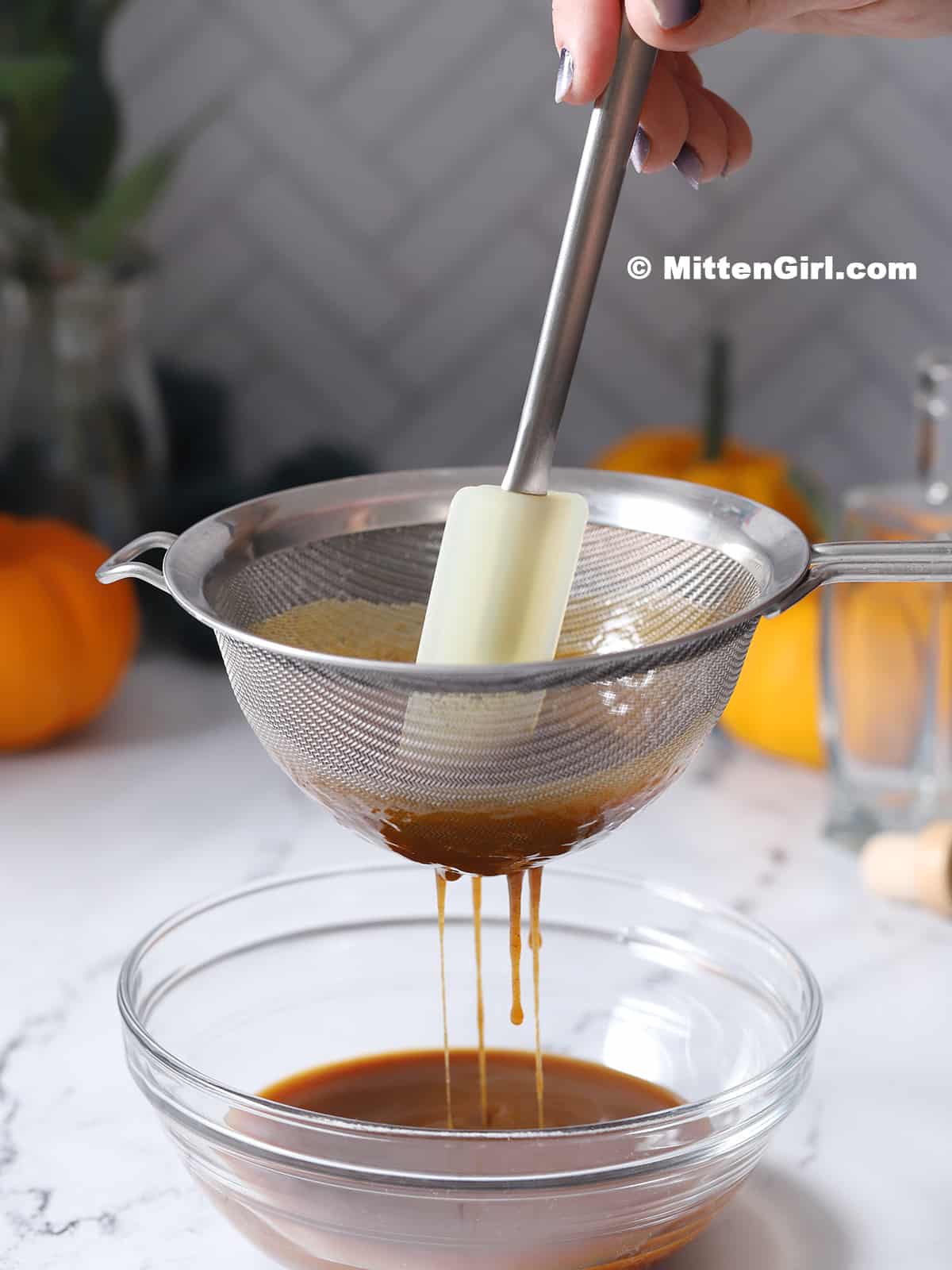 A hand stirring a spatula in a strainer of syrup held over a bowl.