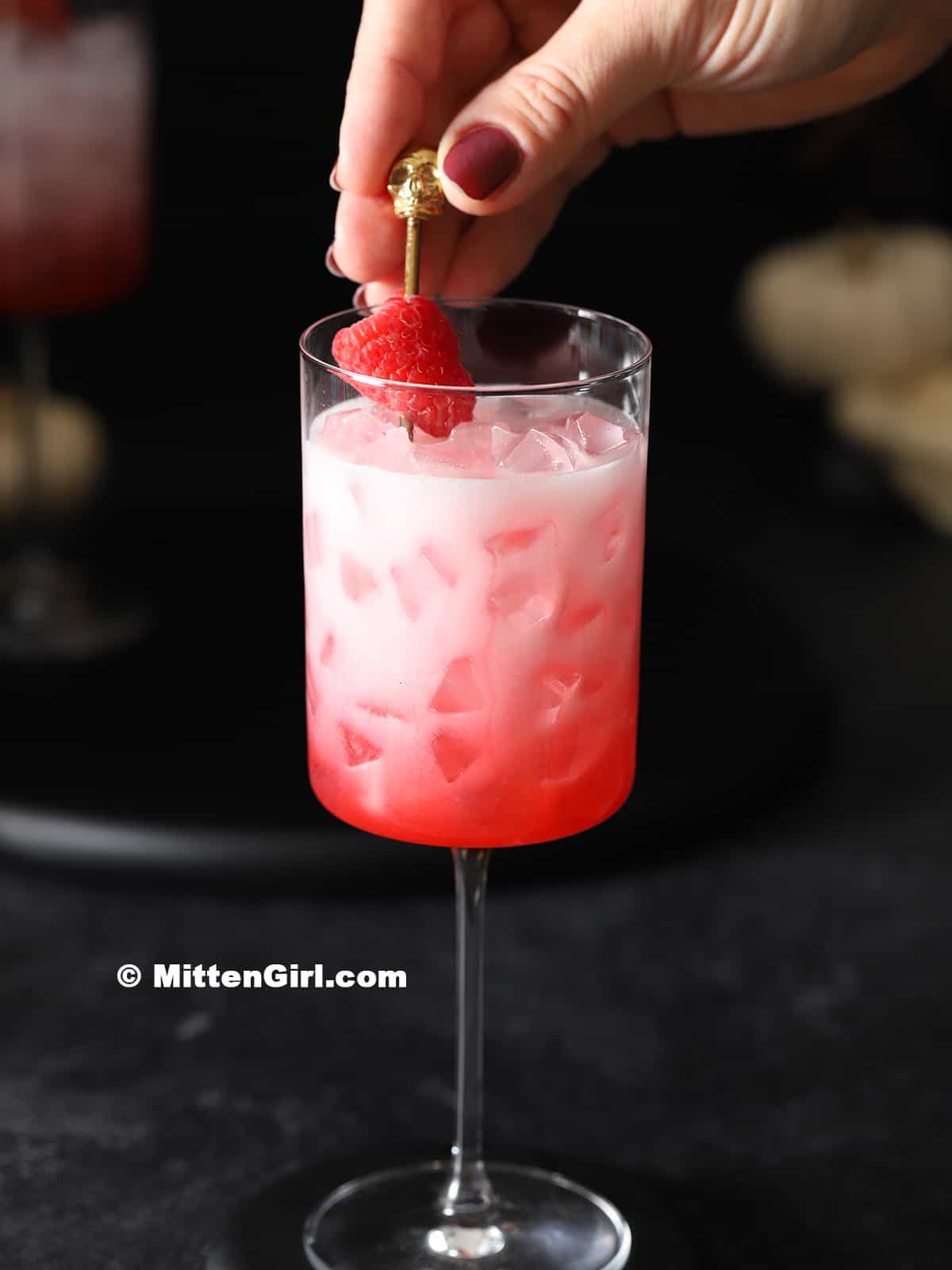 A hand placing a cocktail pick with fresh raspberries into a bloody ghost cocktail.