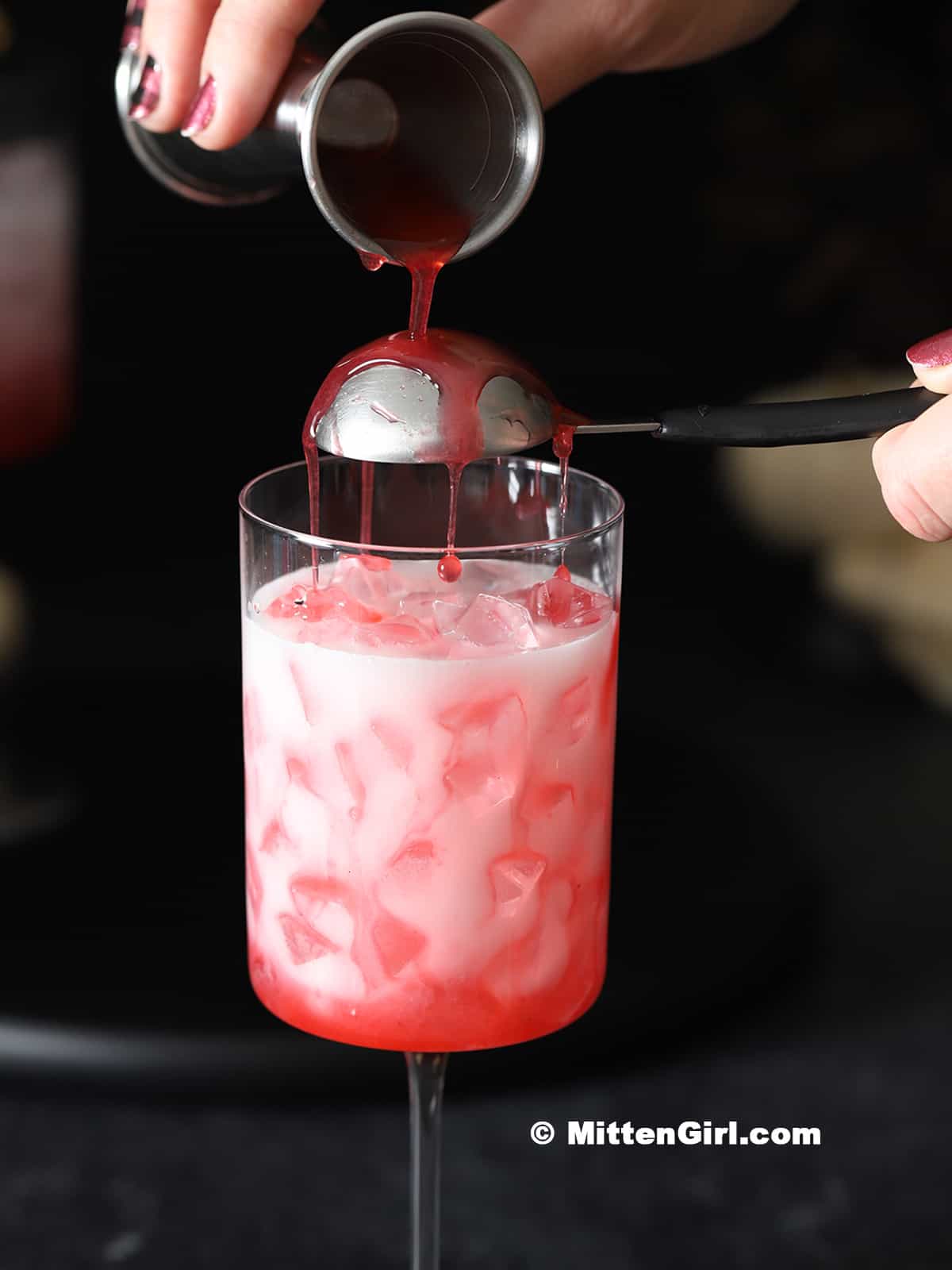 Raspberry syrup being poured over the back of a tablespoon held over a glass.