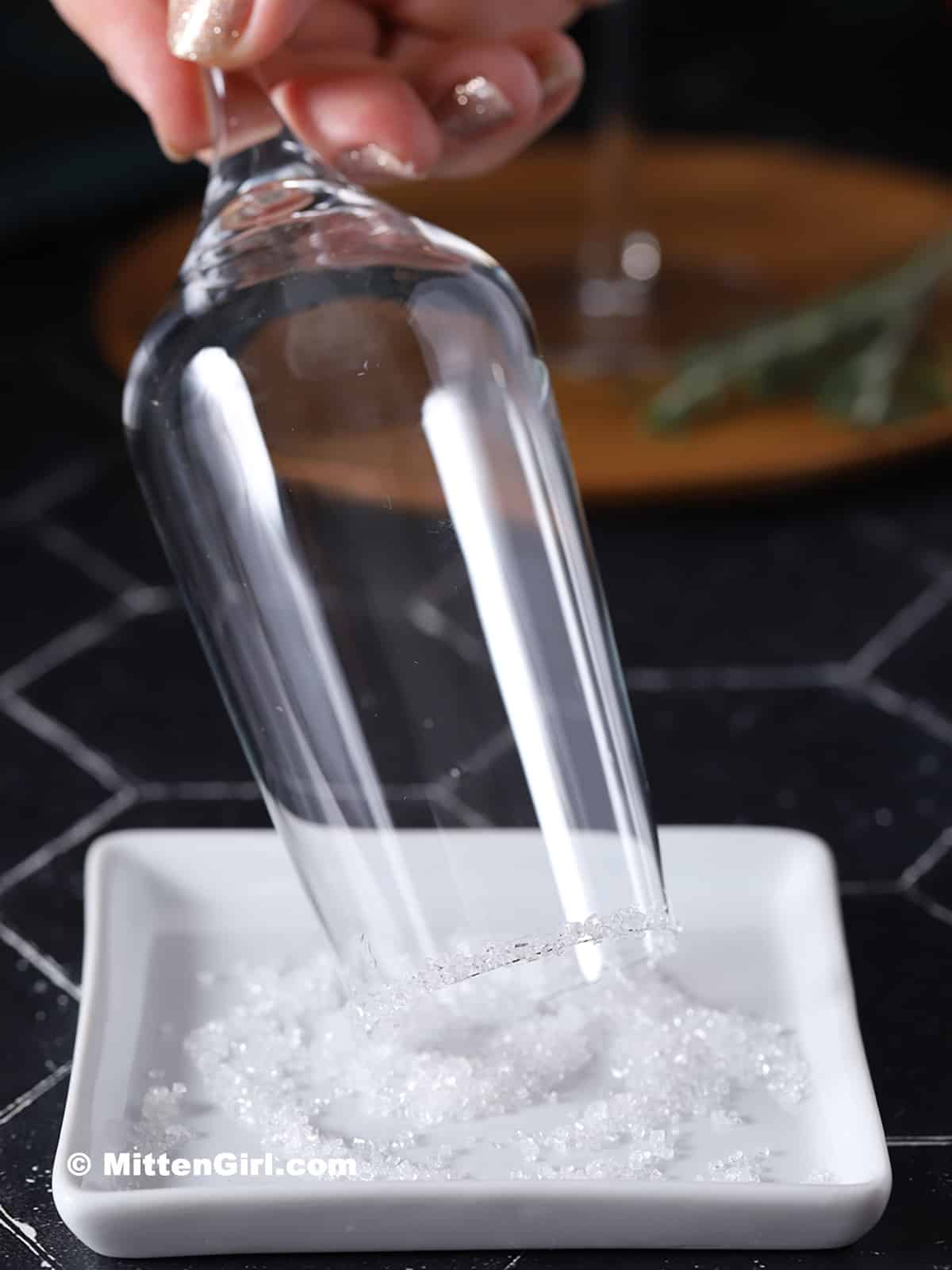 A hand dipping a champagne flute into a dish of decorating sugar.
