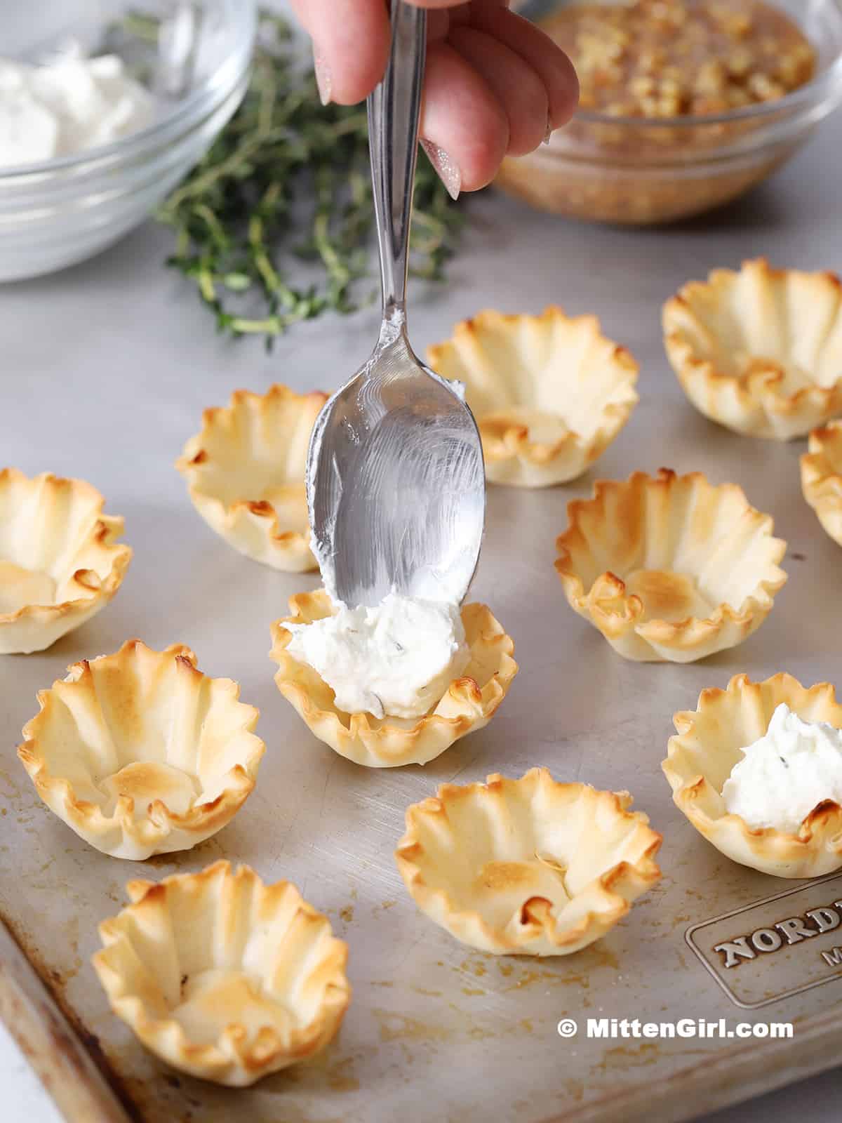 A hand placing a spoon with goat cheese mixture into a phyllo cup.
