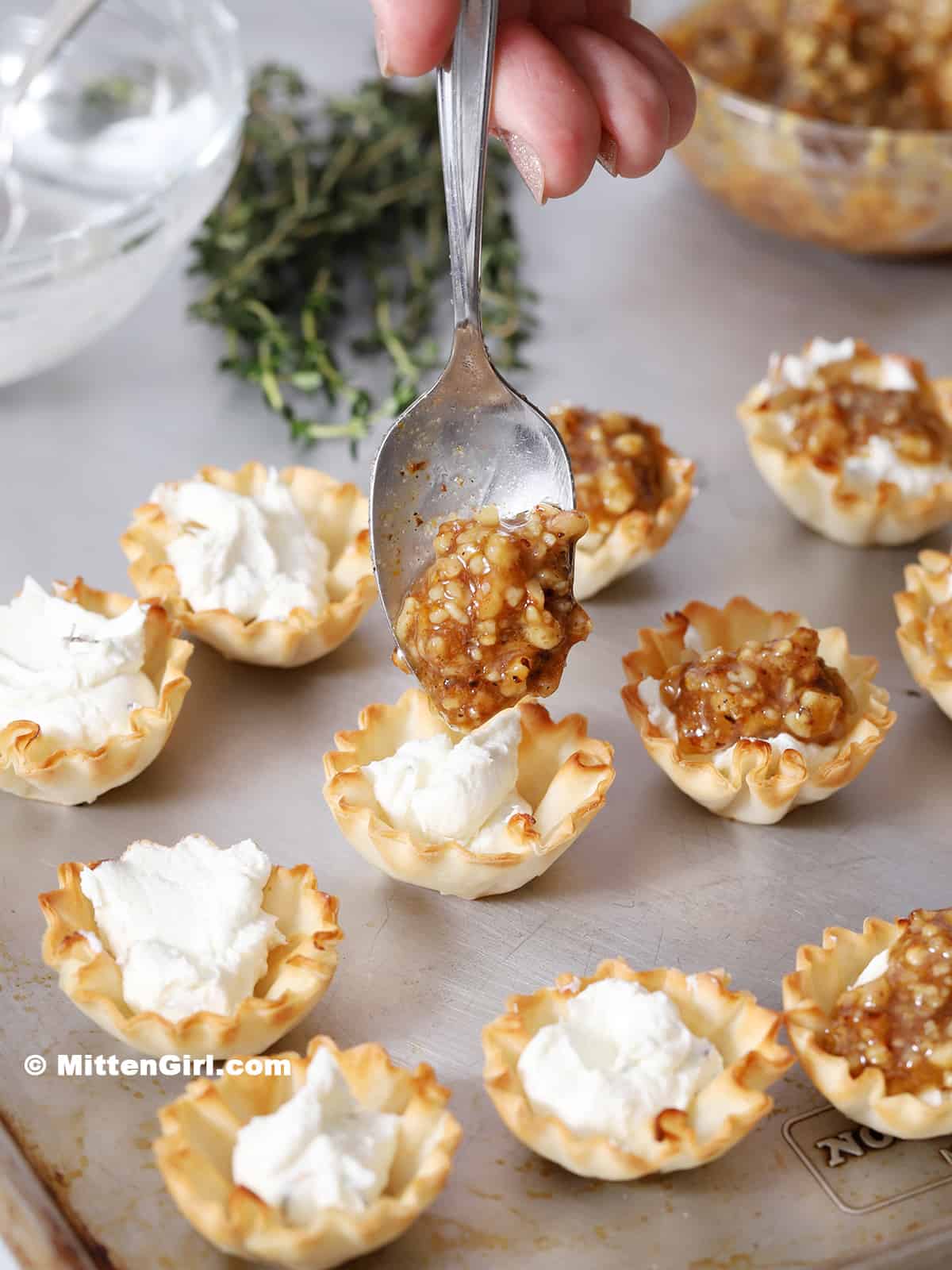 A hand placing a spoon of walnut and honey topping onto a filled phyllo cup.