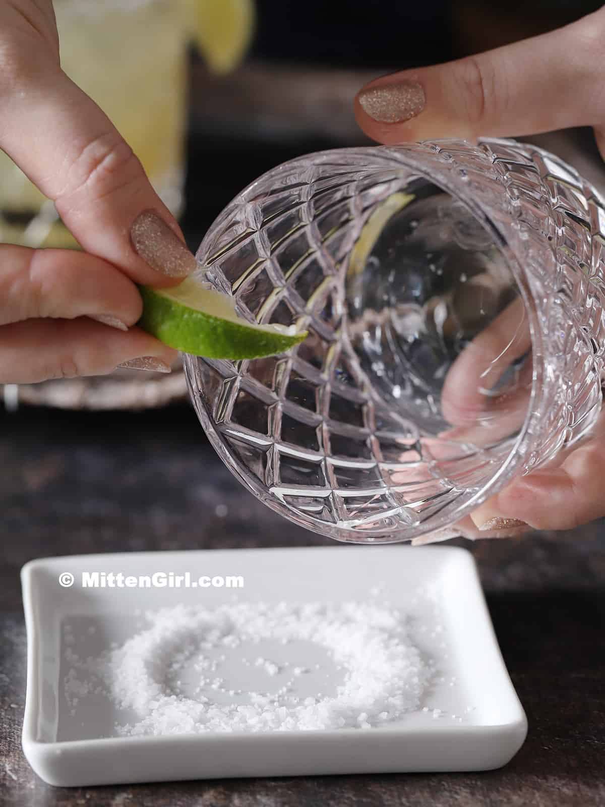 A hand running a lime slice around the rim of a rocks glass.