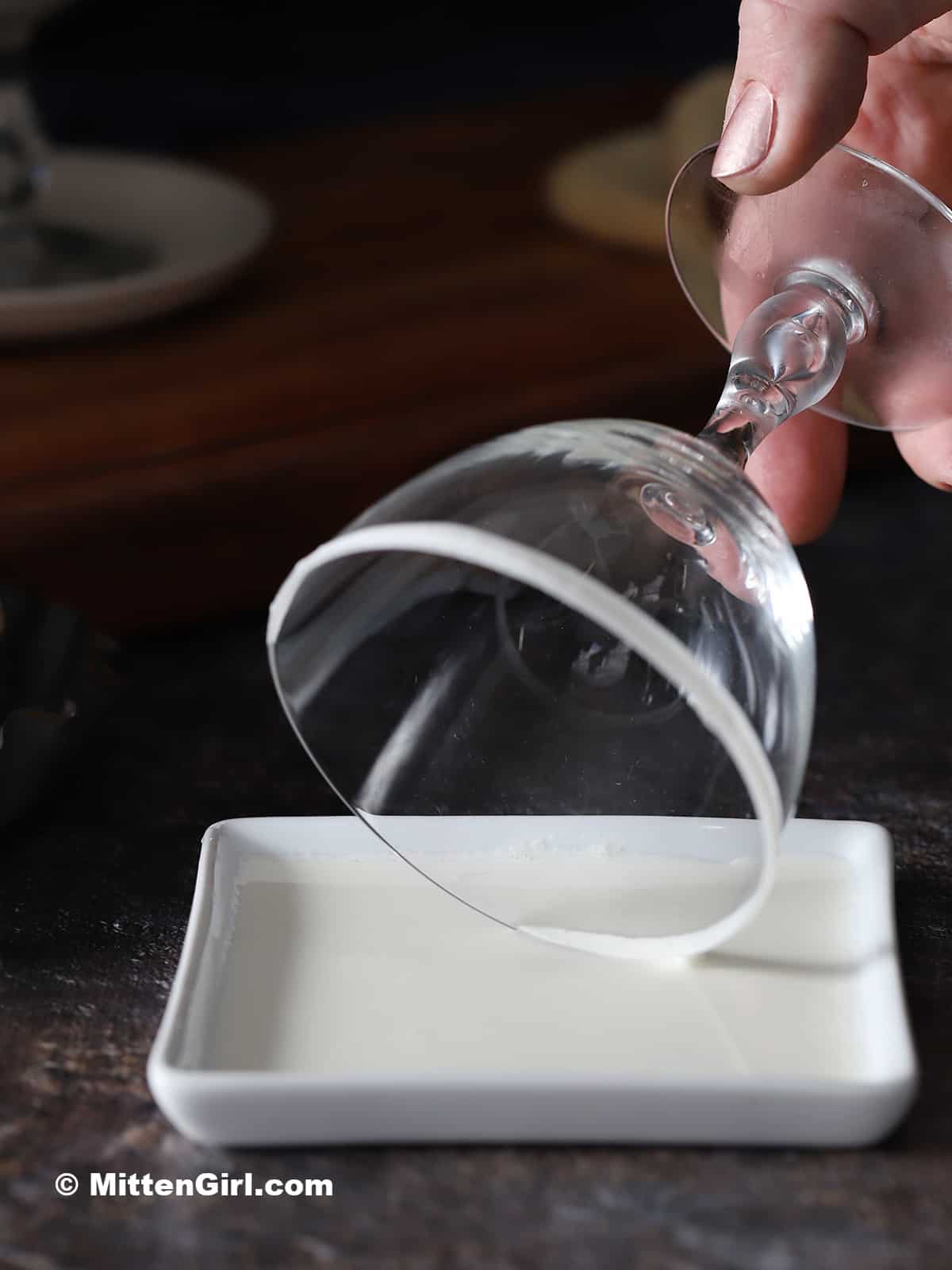 A hand dipping the rim of a glass into a small plate of heavy cream. 