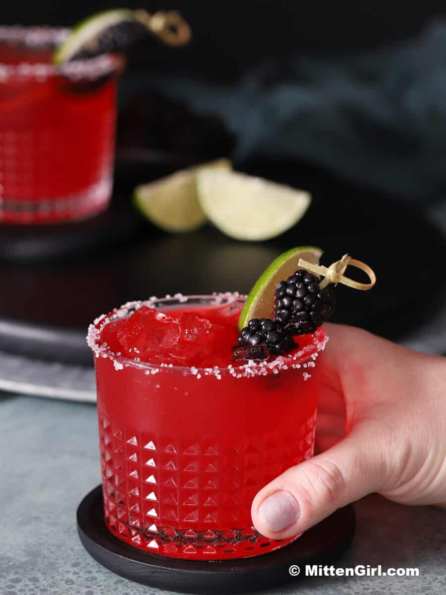 A hand picking up a blackberry margarita garnished with fresh blackberries and lime slices.