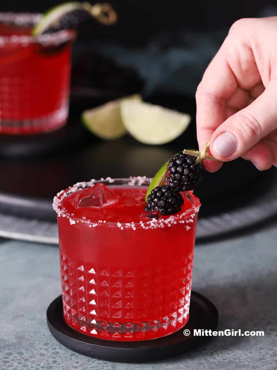 A hand placing a fresh blackberry garnish on a margarita.