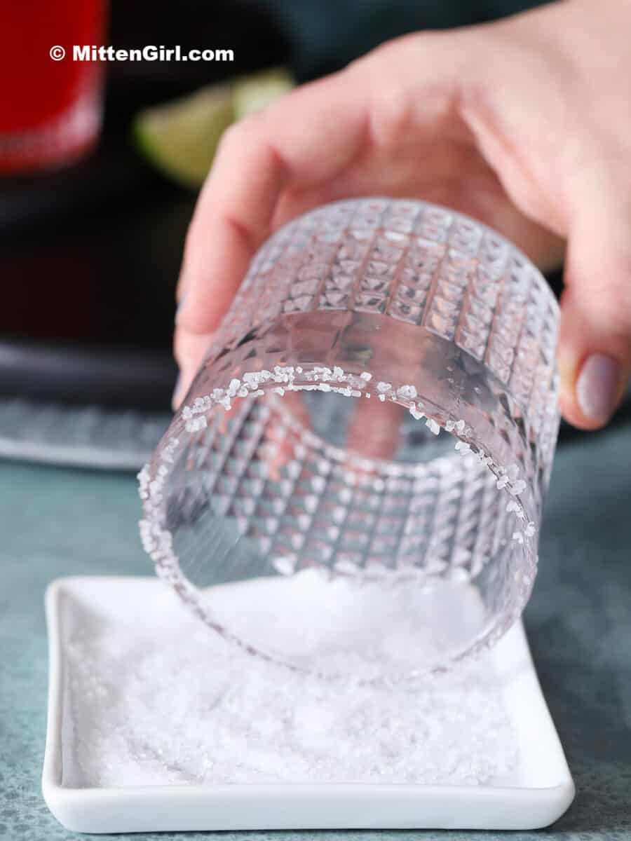 A hand dipping the rim of a glass into coarse salt.