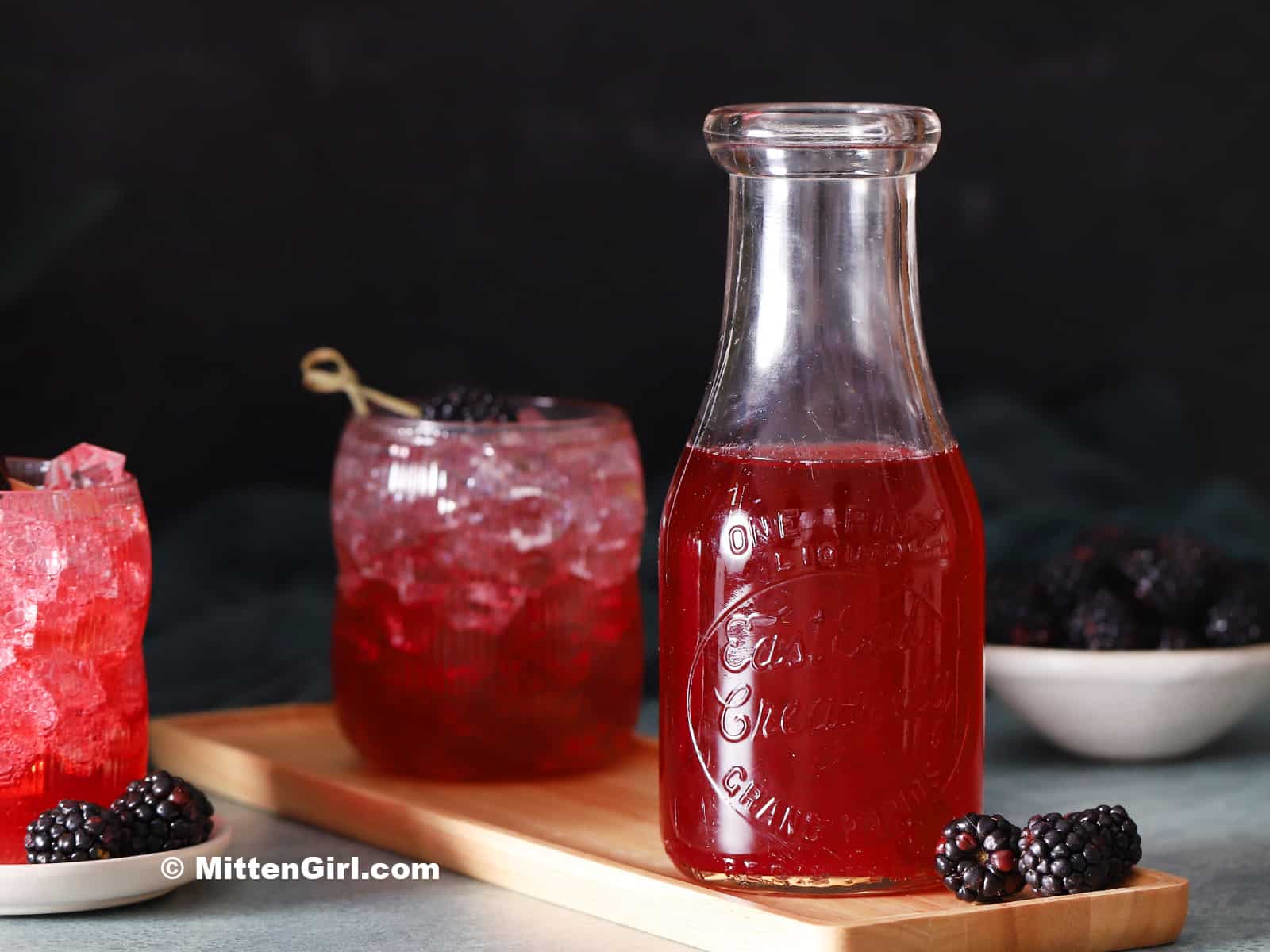 A glass bottle of blackberry syrup next to two glasses of blackberry Italian Sodas.