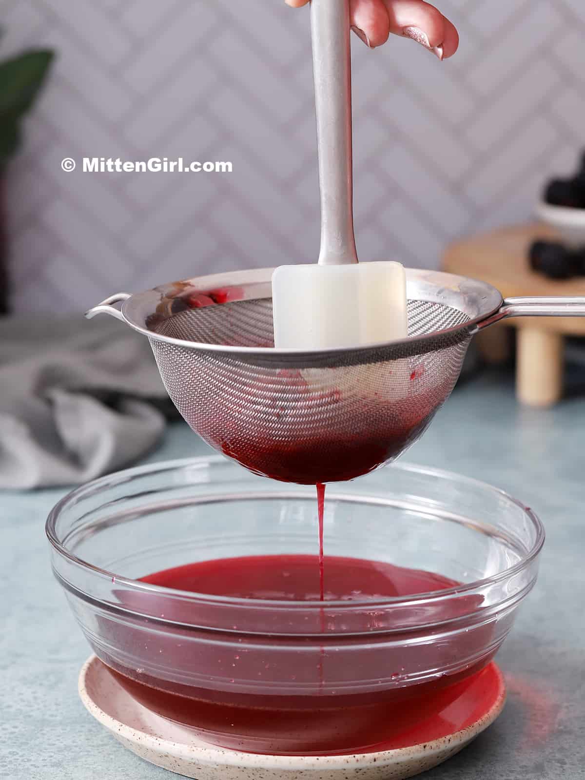 A hand moving a spatula through blackberry syrup in a fine mesh strainer held over a glass bowl. 