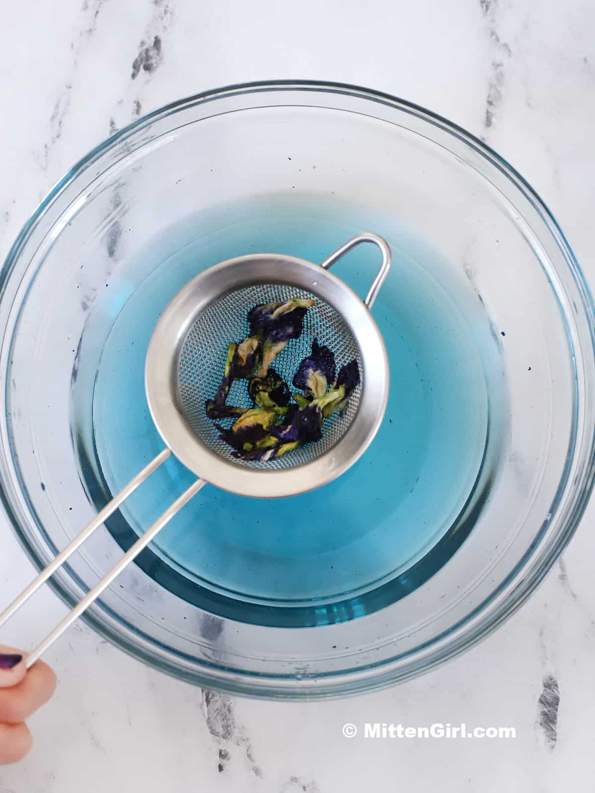 A fine mesh strainer holding the butterfly pea flowers over the finished syrup.