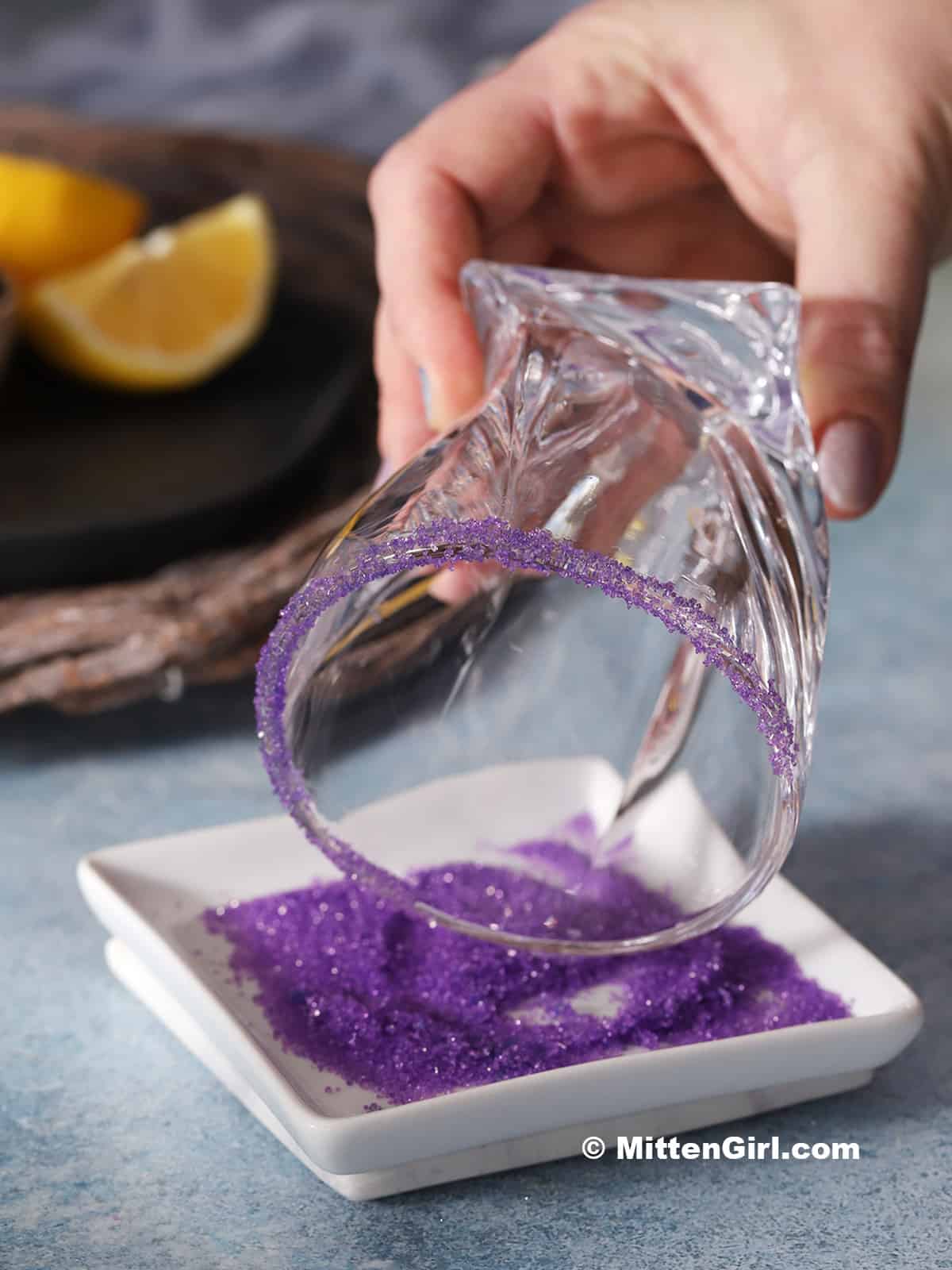 A hand rolling the rim of a glass in purple sugar.