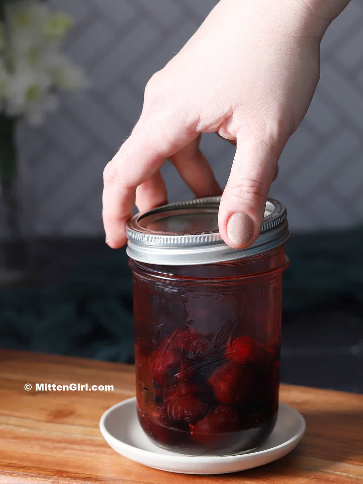 A hand placing a lid on a mason jar filled with cherries and vodka.
