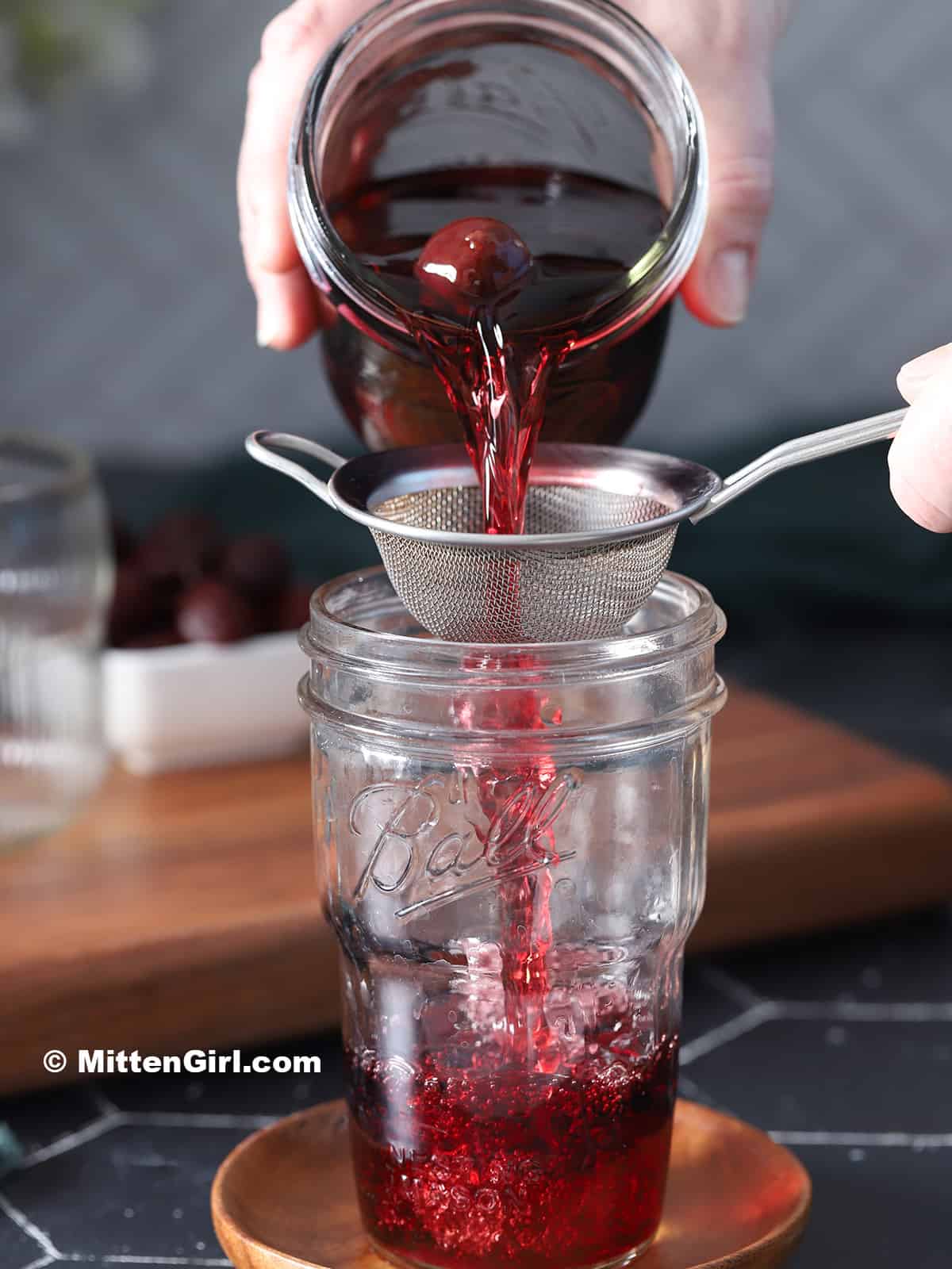 A hand pouring cherry infused vodka through a strainer and into a mason jar.