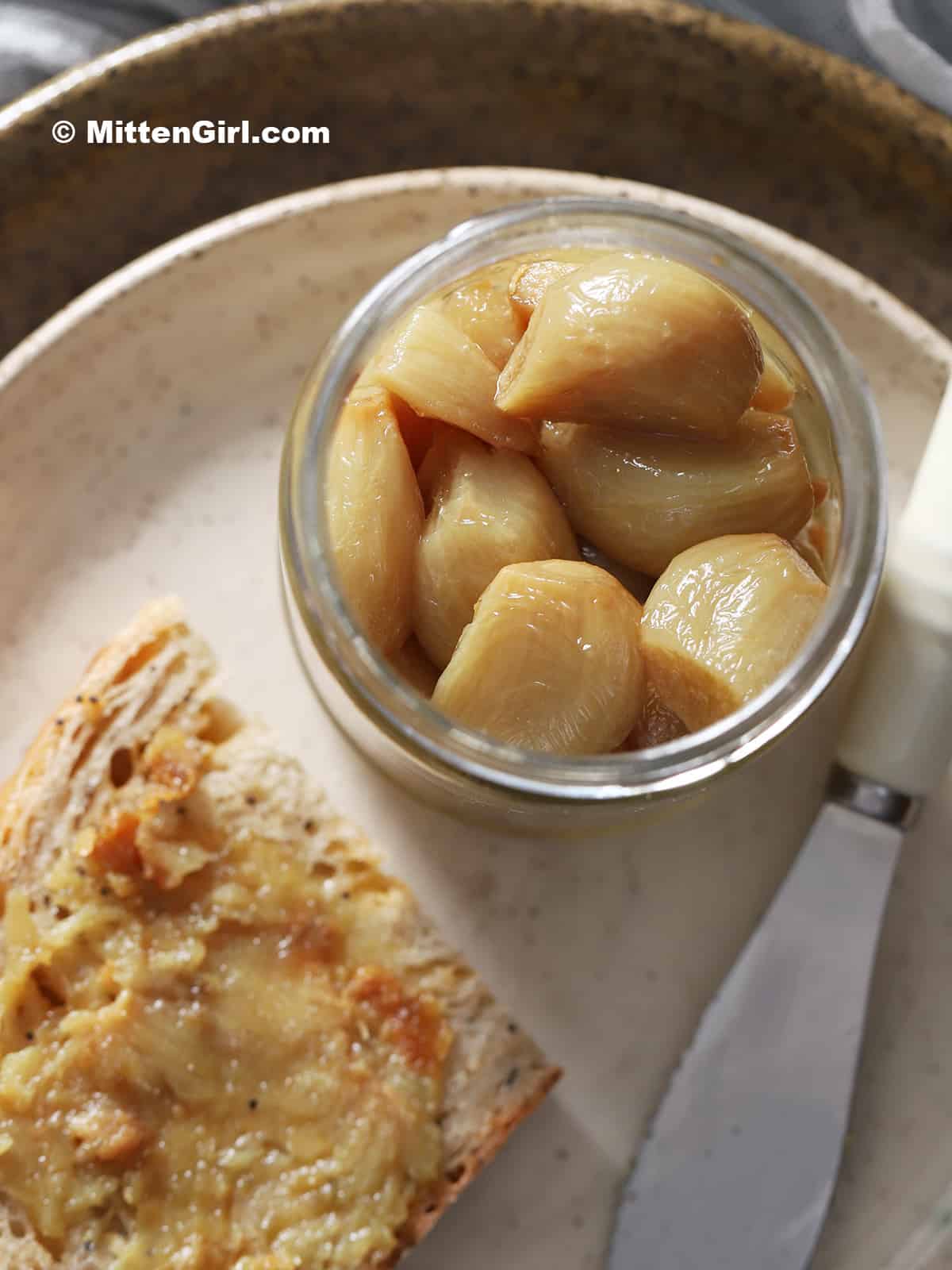 A jar of roasted garlic cloves next to a slice of bread and a spreader.
