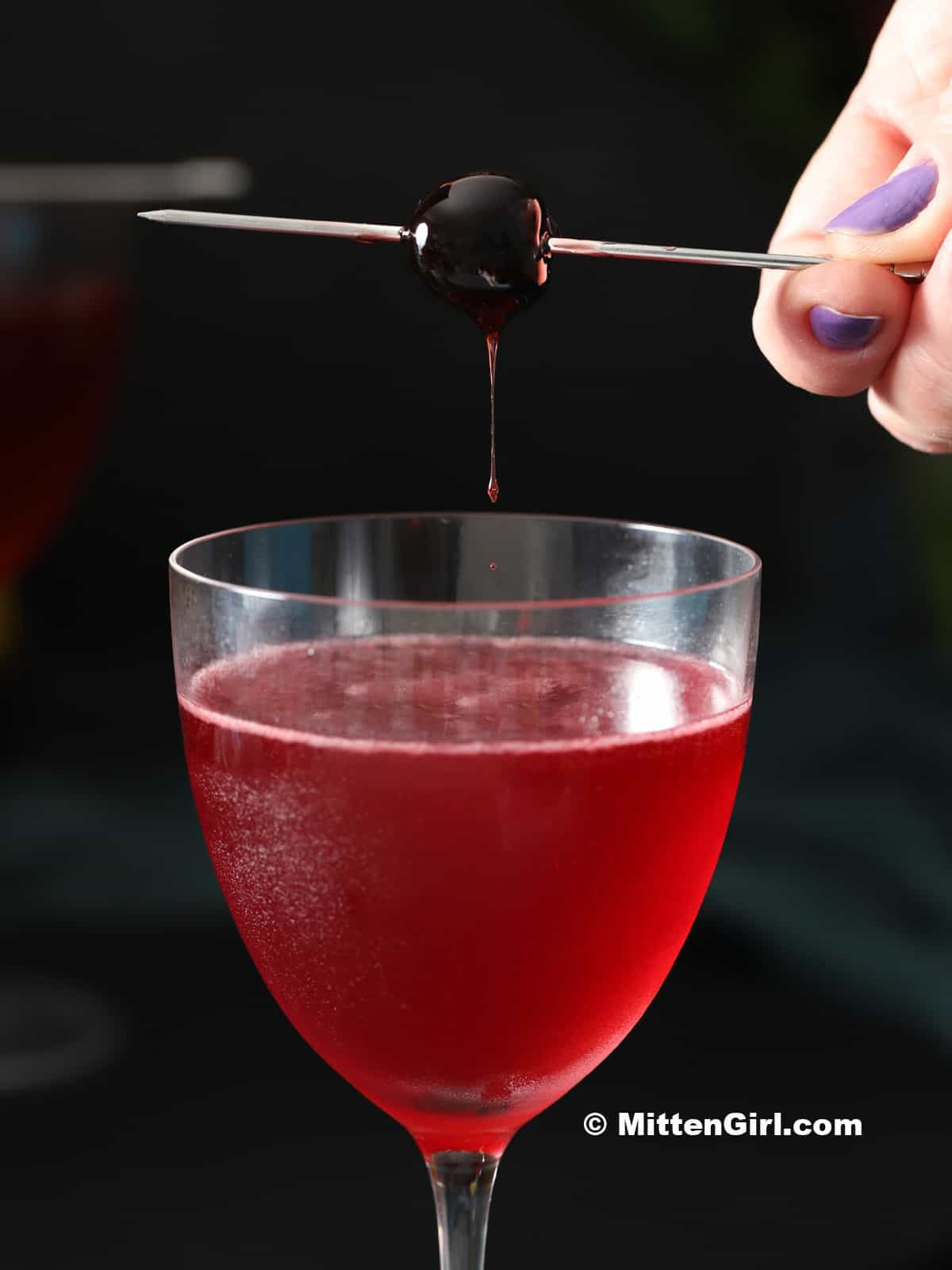 A cocktail pick with a maraschino cherry being set on top of a cocktail glass.