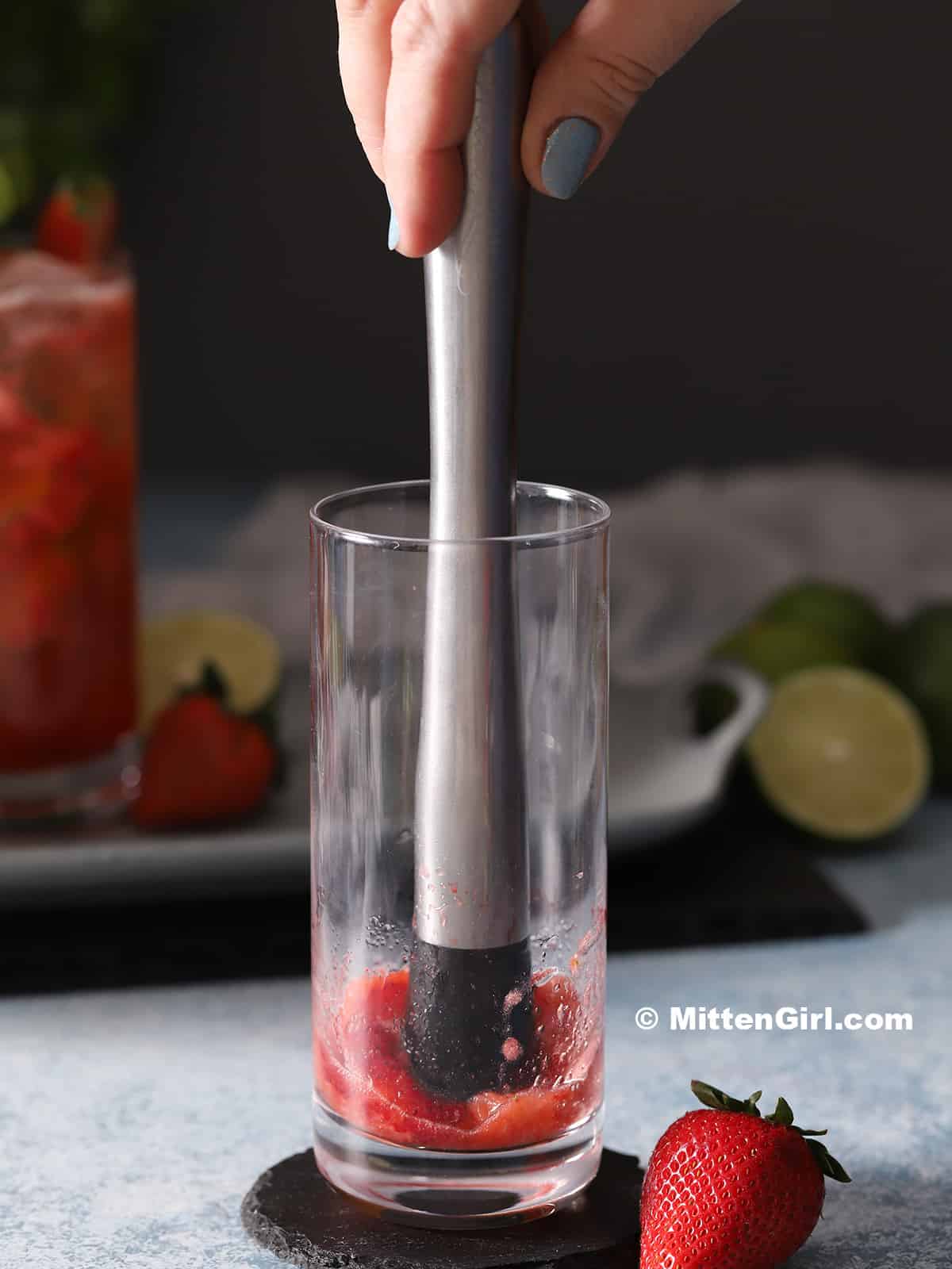 A hand muddling strawberries in a tall glass.