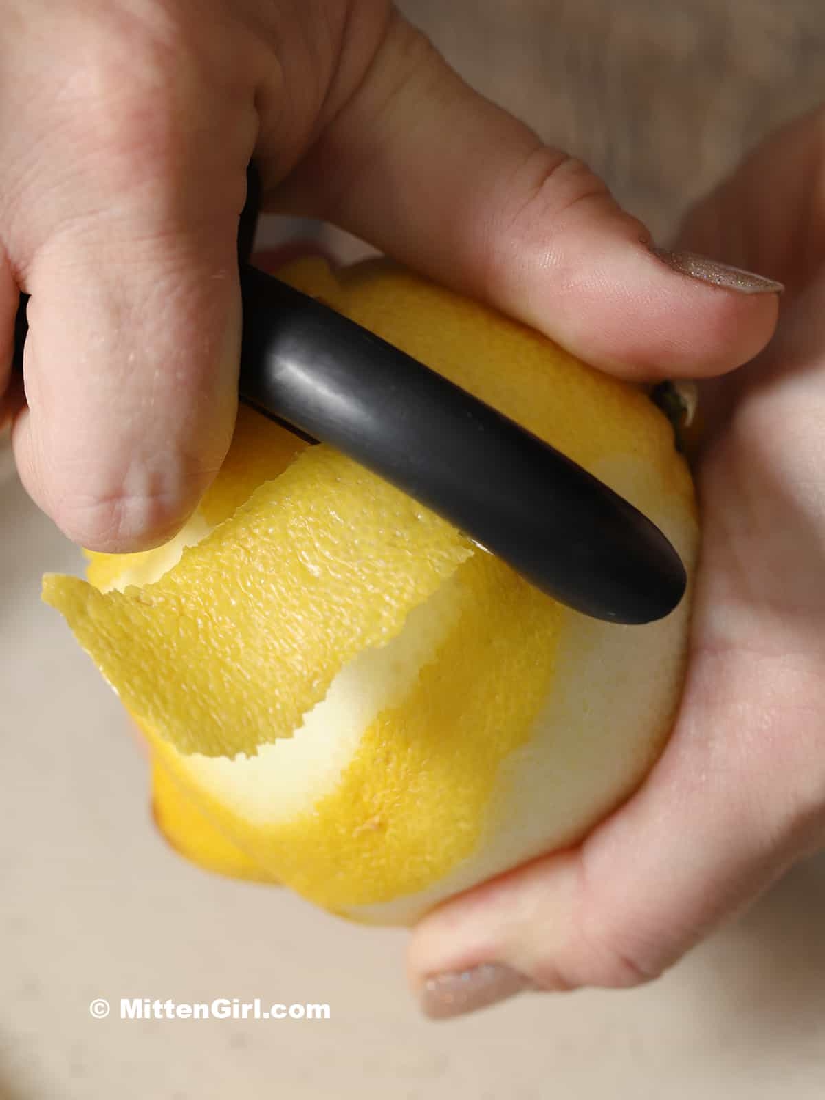 A hand peeling a piece of lemon peel from a fresh lemon. 