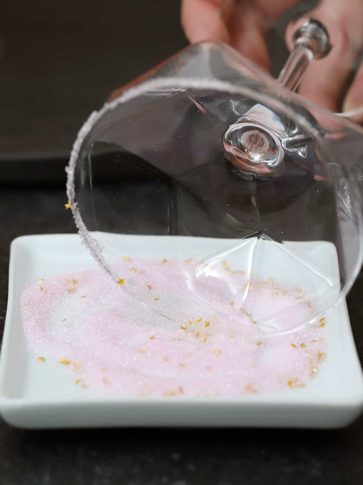 A hand rolling a cocktail glass rim in a dish of pink sugar. 