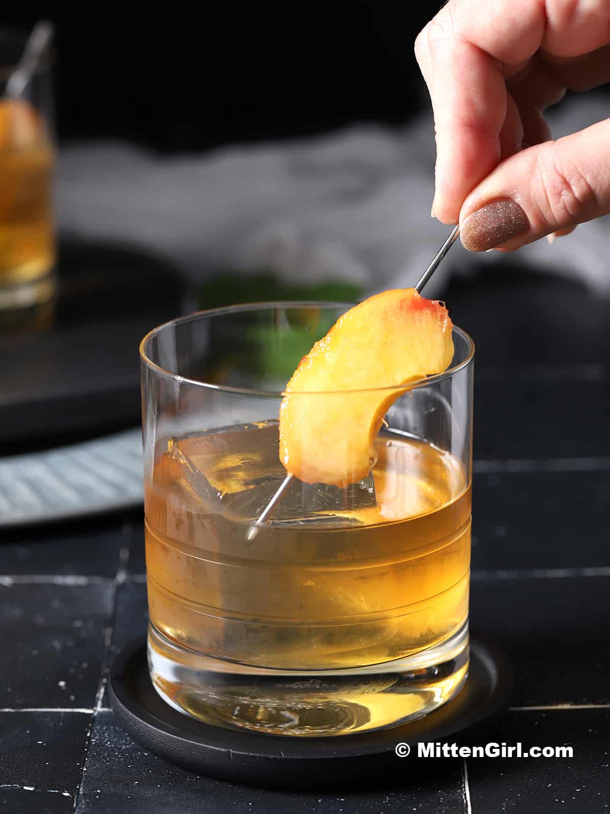 A hand placing a cocktail pick with a slice of peach on it into a glass of peach old fashioned cocktail. 
