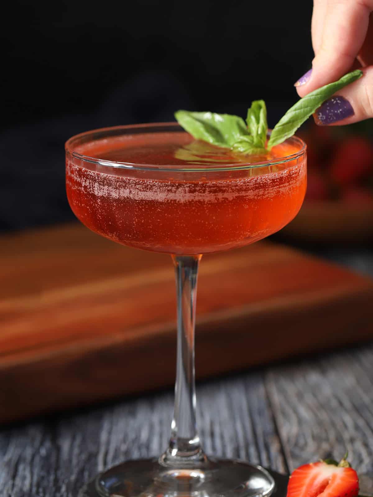A hand garnishing a glass of strawberry basil cocktail with a basil leaf.
