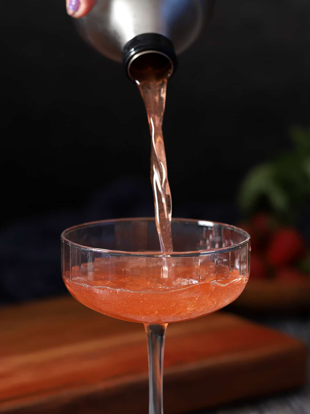 A strawberry basil cocktail being poured from a cocktail shaker into a coupe glass .