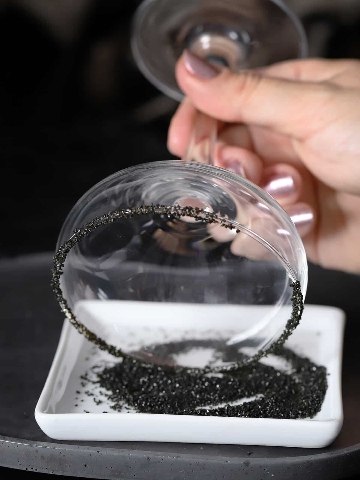 A hand rolling the rim of a glass into black decorating sugar. 